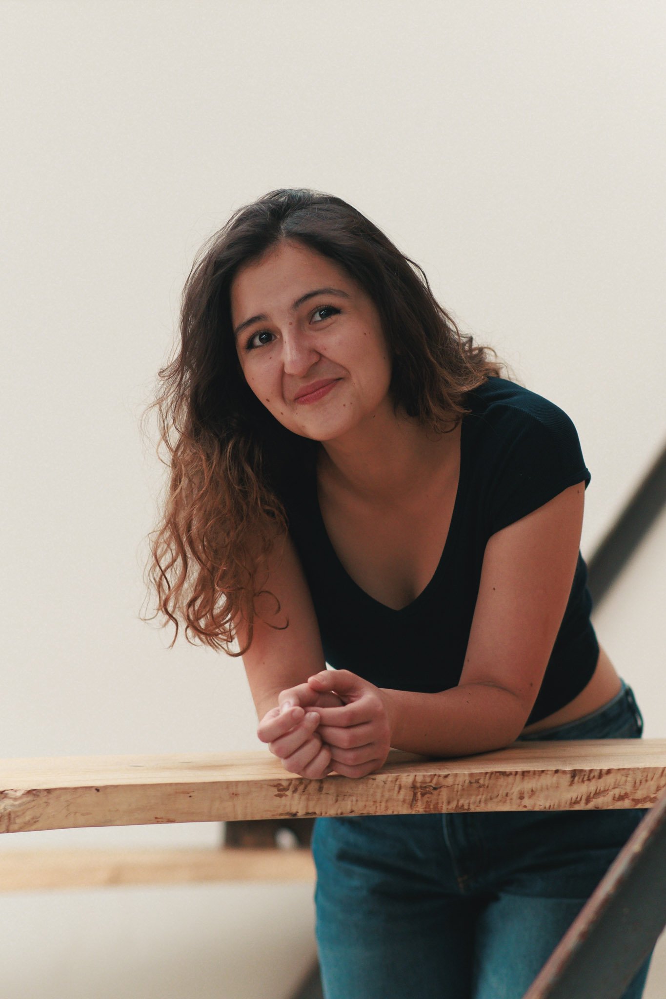 A young woman with wavy brown hair wearing a black top and blue jeans, leaning on a wooden railing, smiling at the camera against a white background.