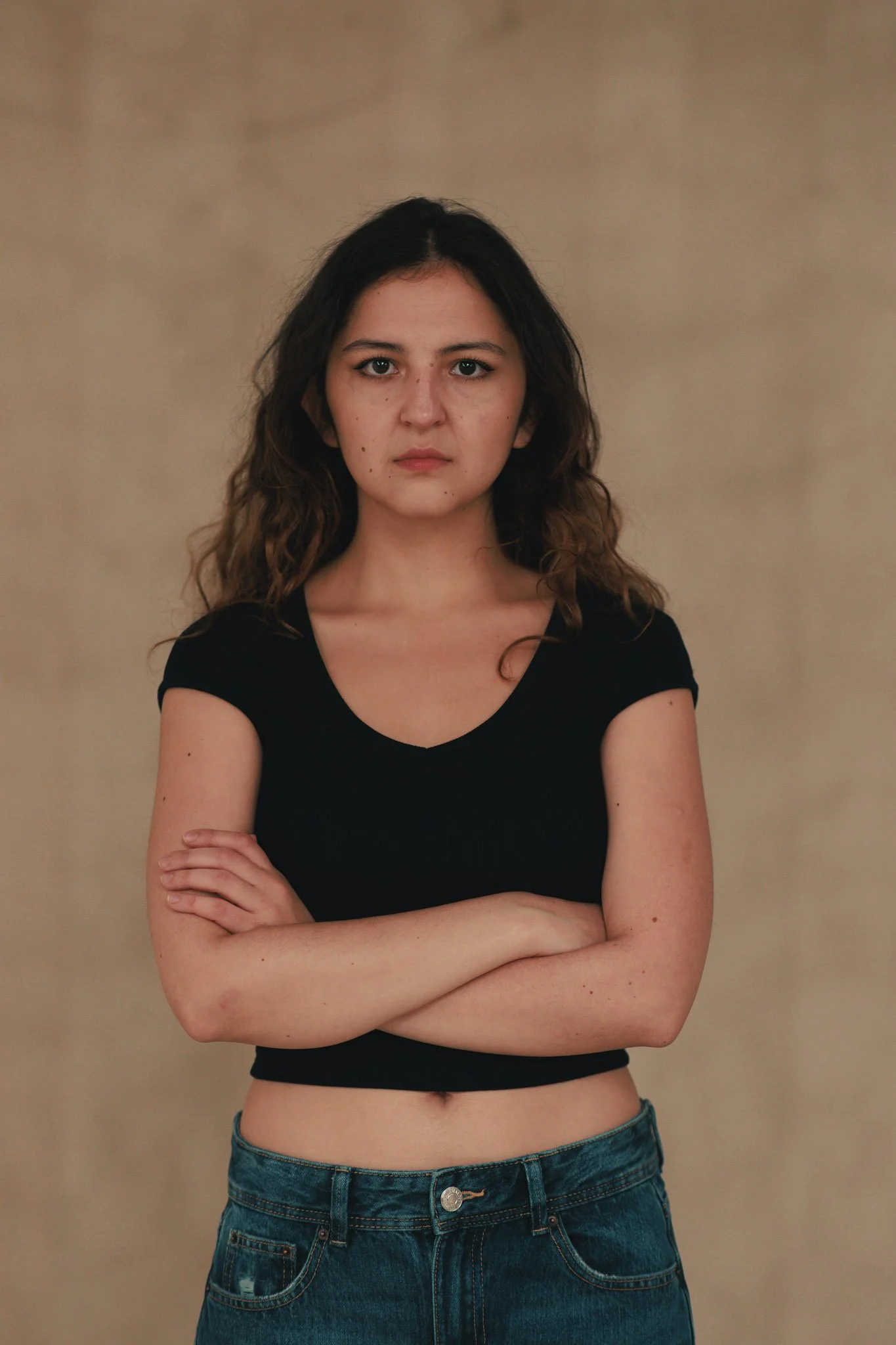 A young woman with wavy brown hair, crossing her arms, wearing a black crop top and blue jeans, standing against a plain beige background.