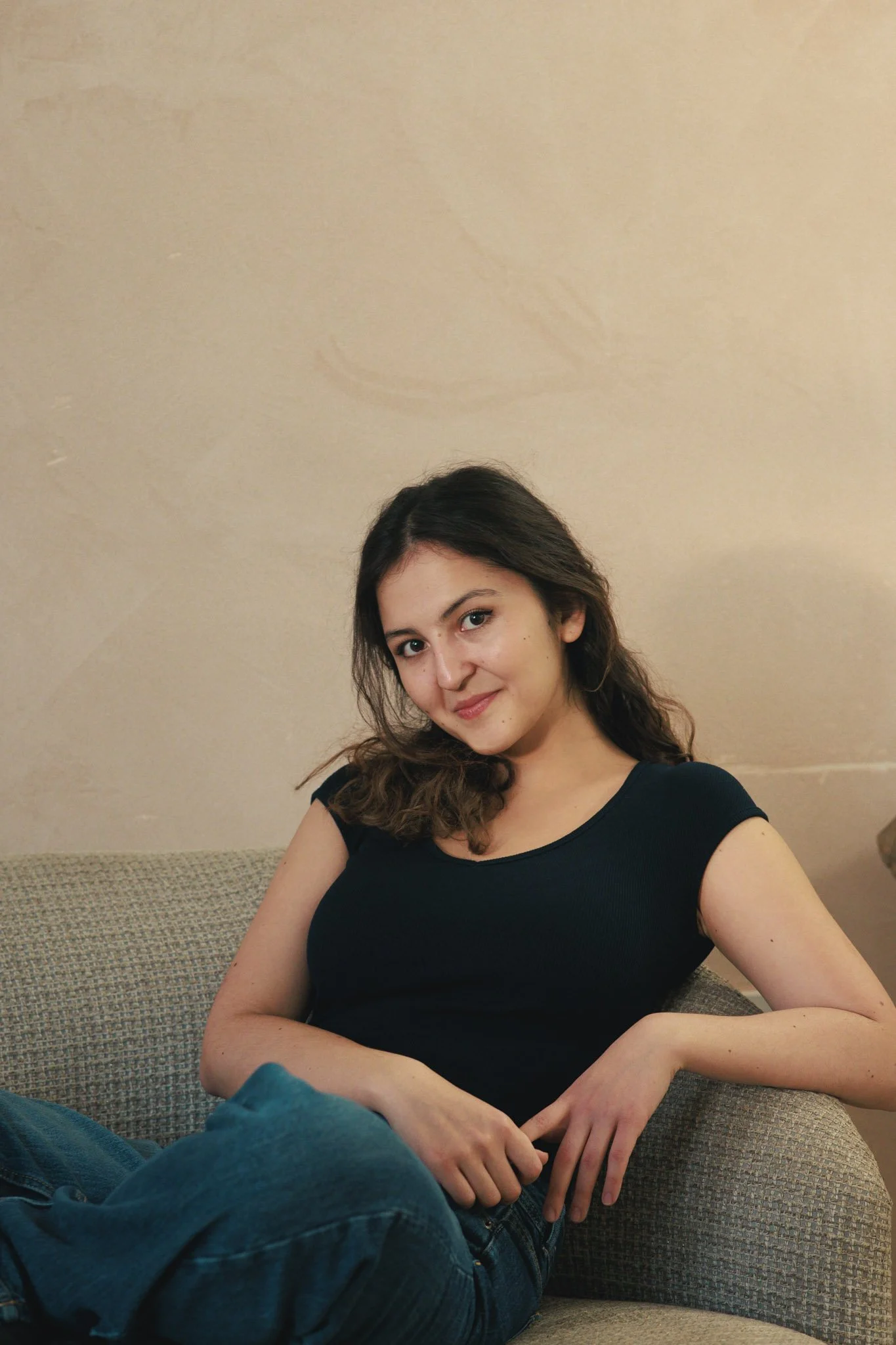 A young woman with dark wavy hair, wearing a black short-sleeve top and blue jeans, sitting on a beige couch against a plain beige wall, smiling gently at the camera.