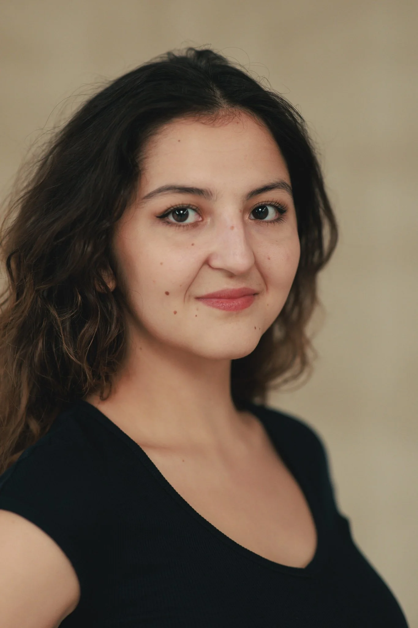 A young woman with shoulder-length, curly brown hair smiling at the camera against a beige background.