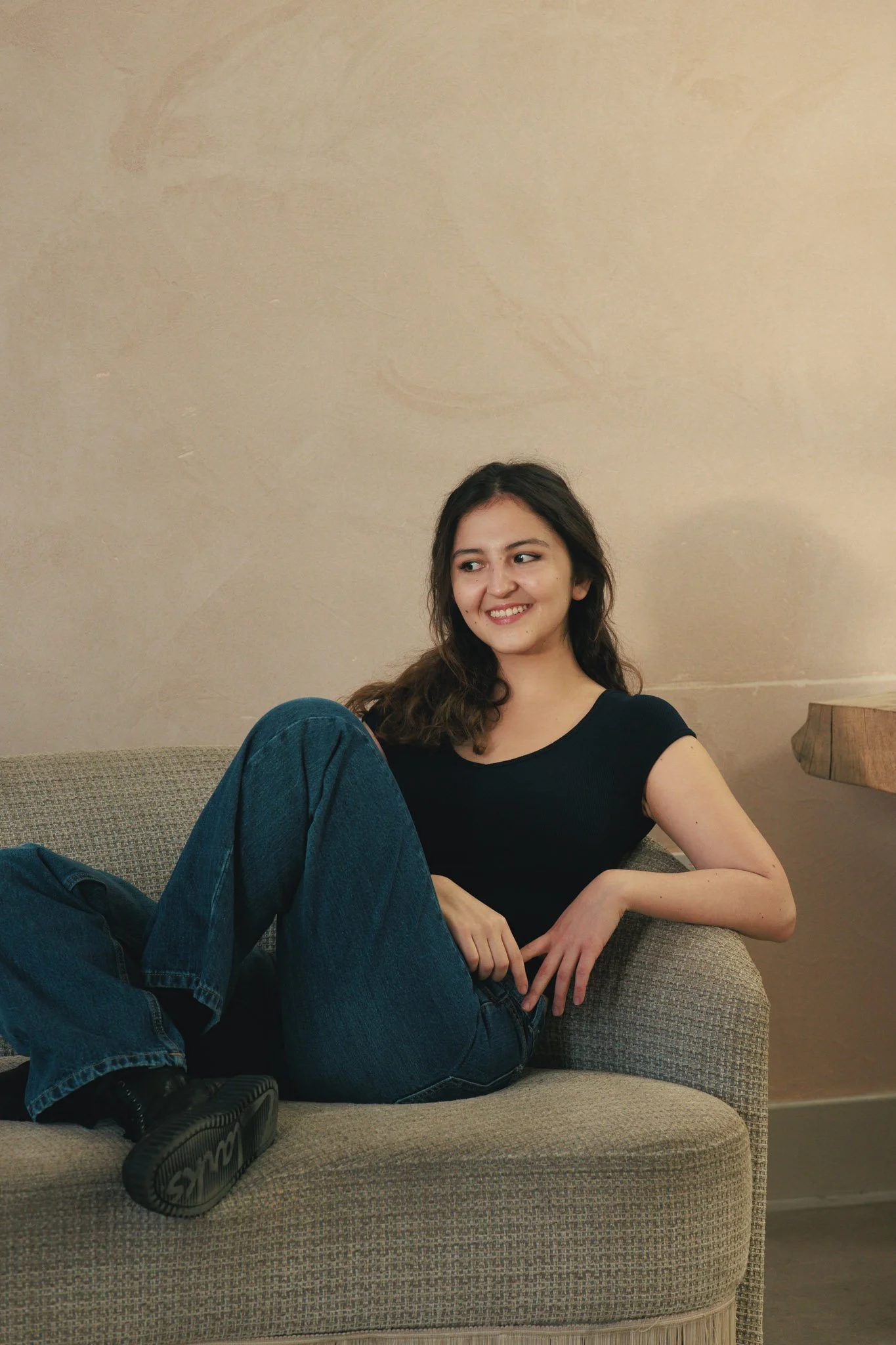 Young woman with long dark hair sitting casually on a couch, smiling and looking to her left. She is wearing a black t-shirt, blue jeans, and black shoes. The background features a plain beige wall and wooden furniture.