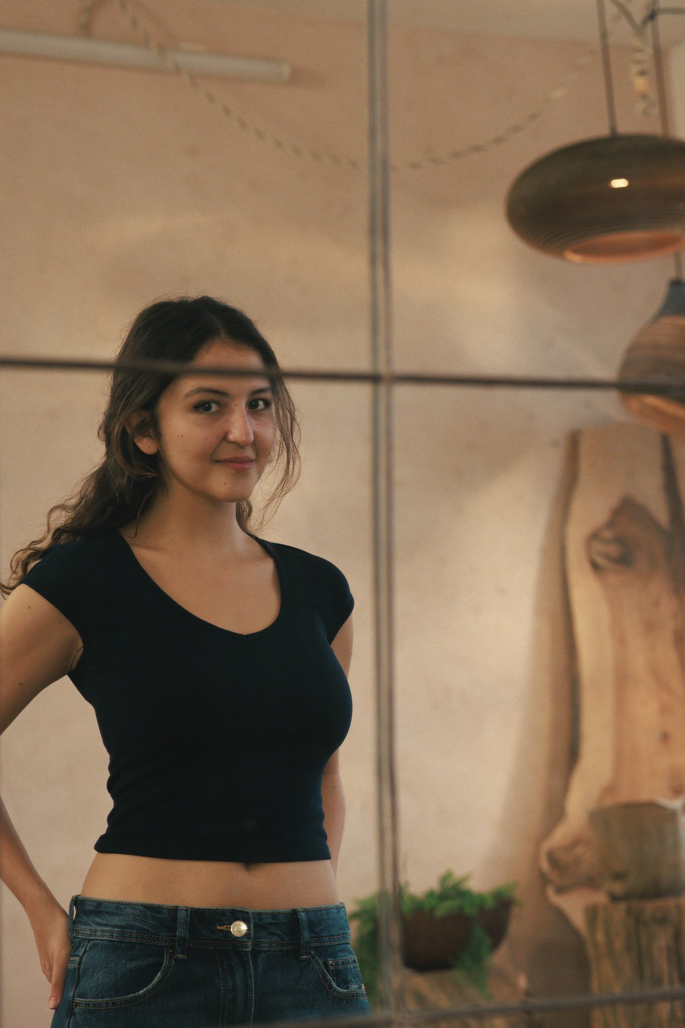 Young woman with long wavy hair looking at the camera, standing indoors behind a glass mirror.