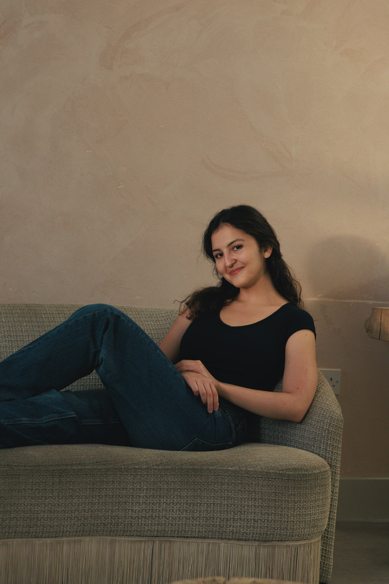 A young woman with dark hair relaxing on a beige sofa, wearing a black t-shirt and blue jeans, smiling at the camera in a cozy room.