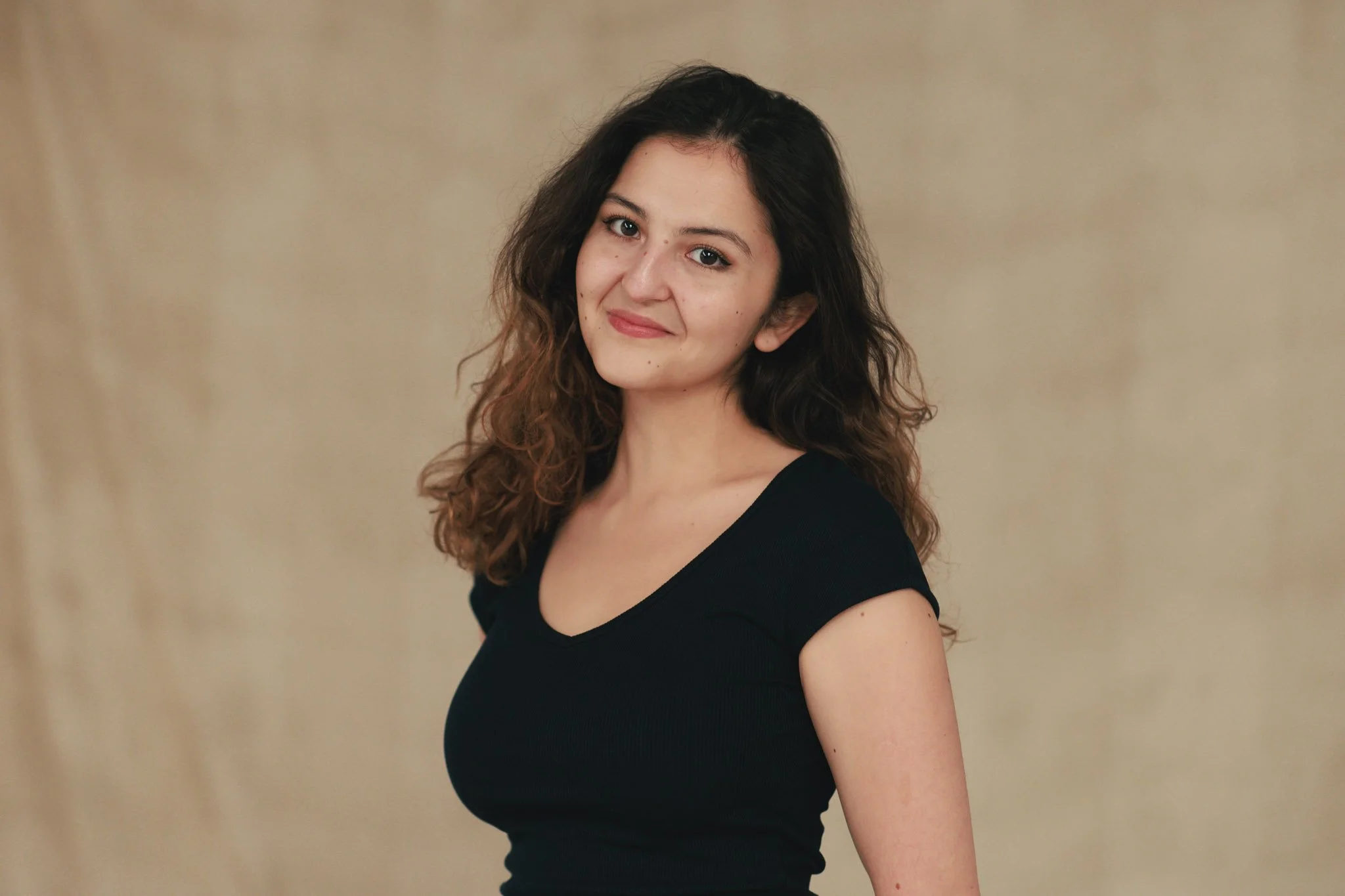 A young woman with long, curly brown hair wearing a black V-neck shirt, smiling softly and standing against a beige background.