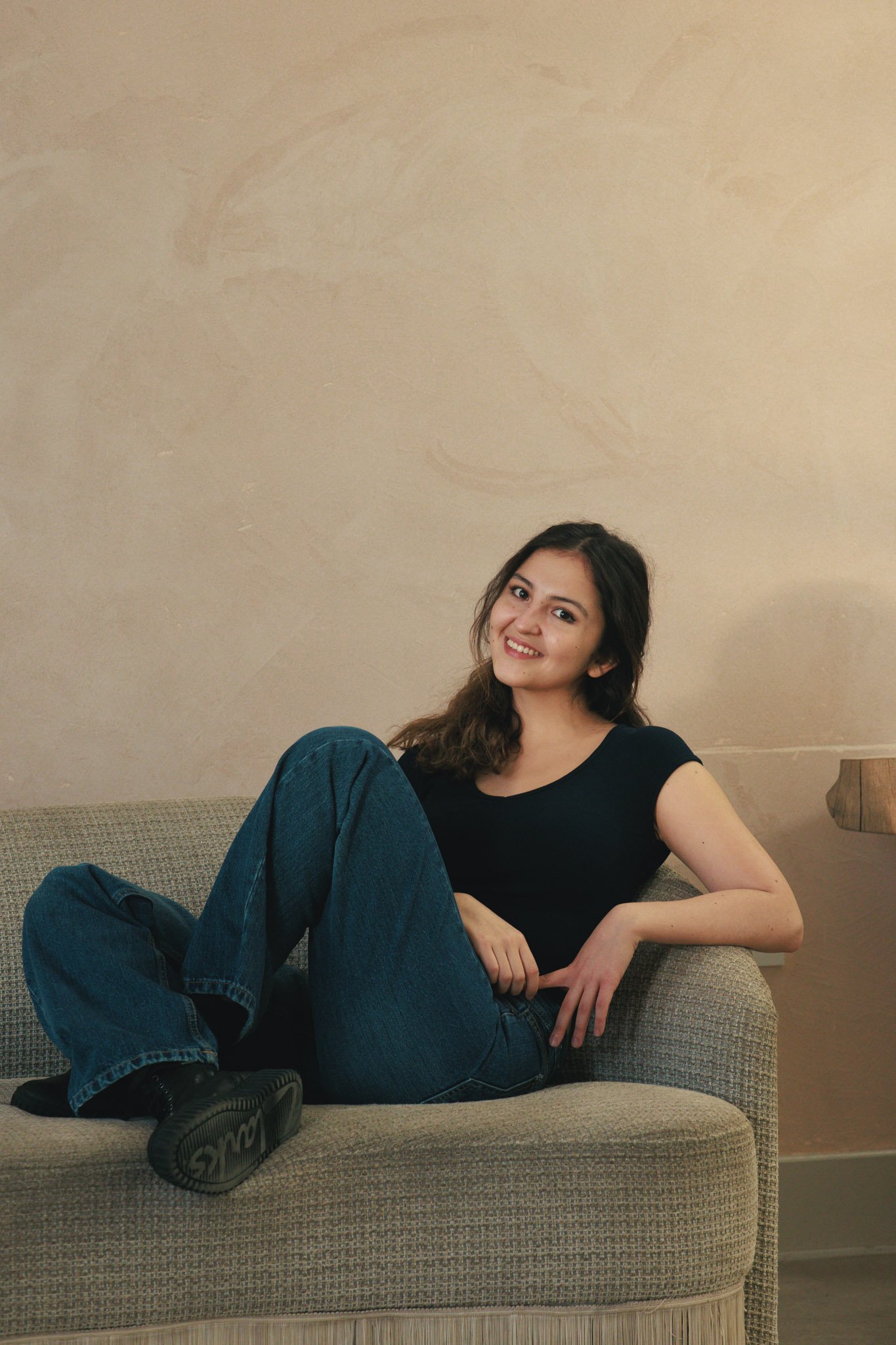 A young woman with long brown hair, wearing a black t-shirt and blue jeans, is sitting comfortably on a beige sofa, smiling at the camera.