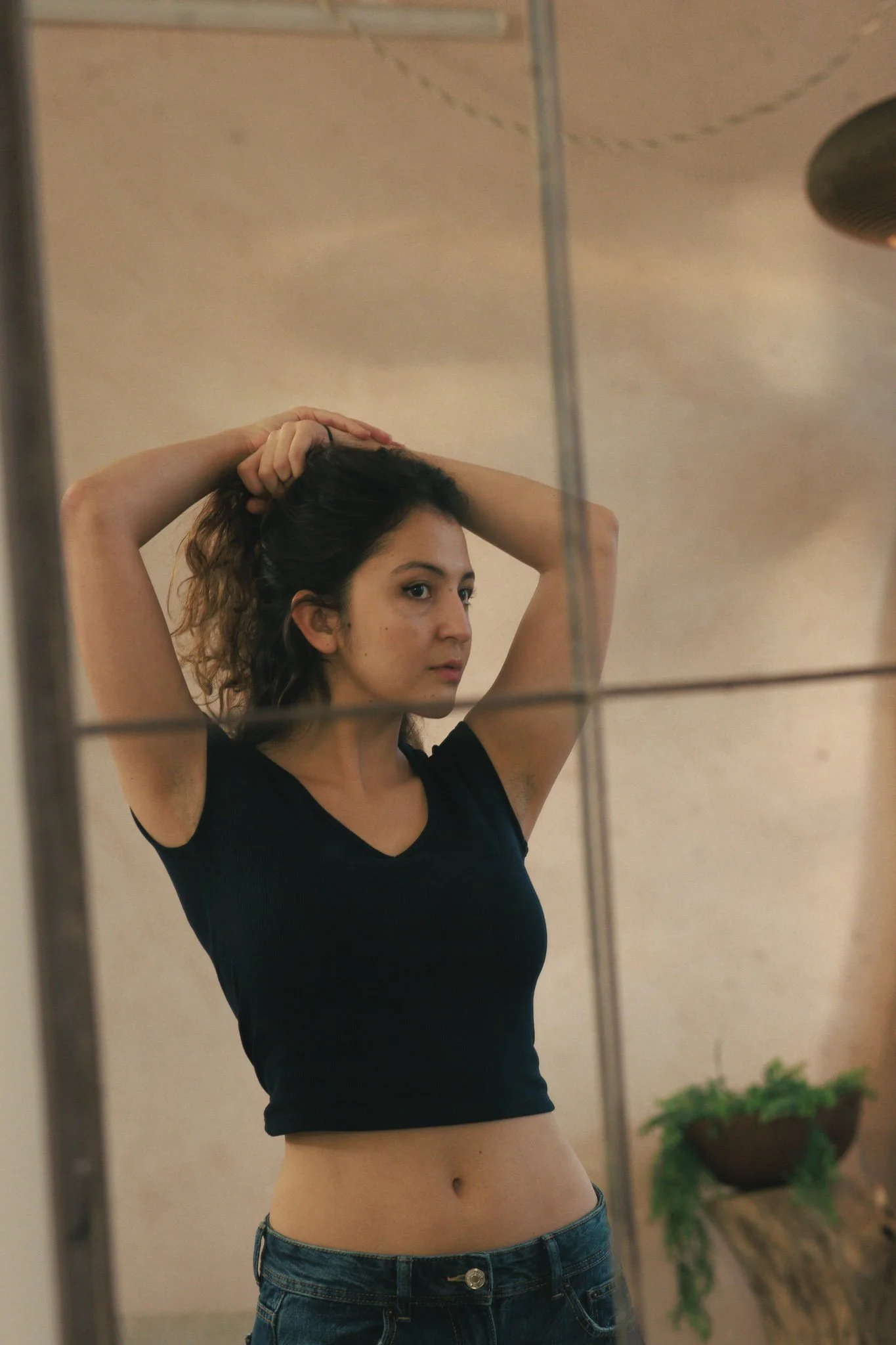 A young woman with dark, wavy hair standing in front of a mirror, wearing a black crop top and blue jeans, touching her hair with both hands.
