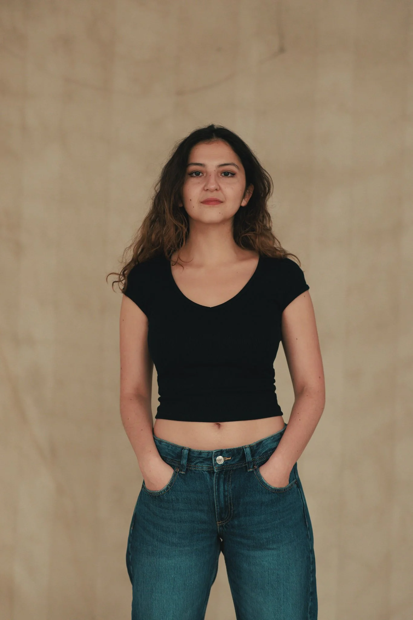 A young woman with long, wavy brown hair, wearing a black short-sleeve crop top and blue jeans, standing with her hands in her pockets against a beige textured background.