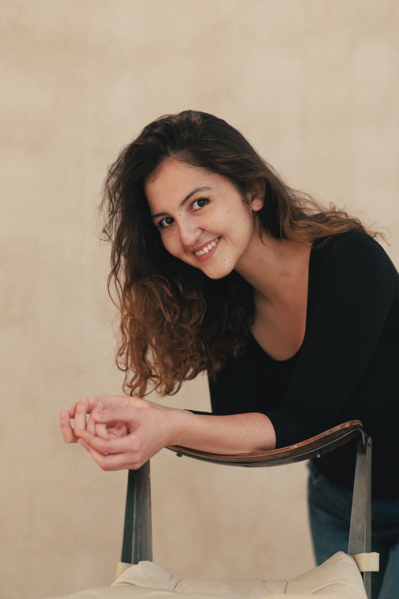 A woman with curly brown hair wearing a black top, smiling and leaning on a chair with a beige cushion against a plain beige wall.