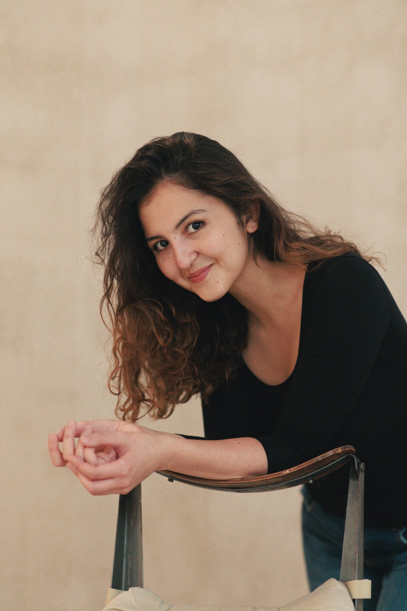 A young woman with brown curly hair wearing a black top, smiling and leaning on a chair with a beige background.