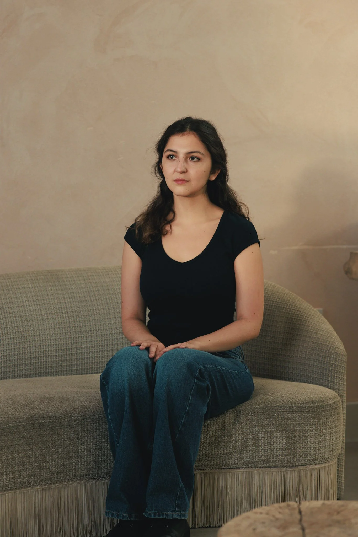 A woman with long dark hair, wearing a black t-shirt and blue jeans, sitting on a green sofa with a neutral expression, in a room with beige walls.