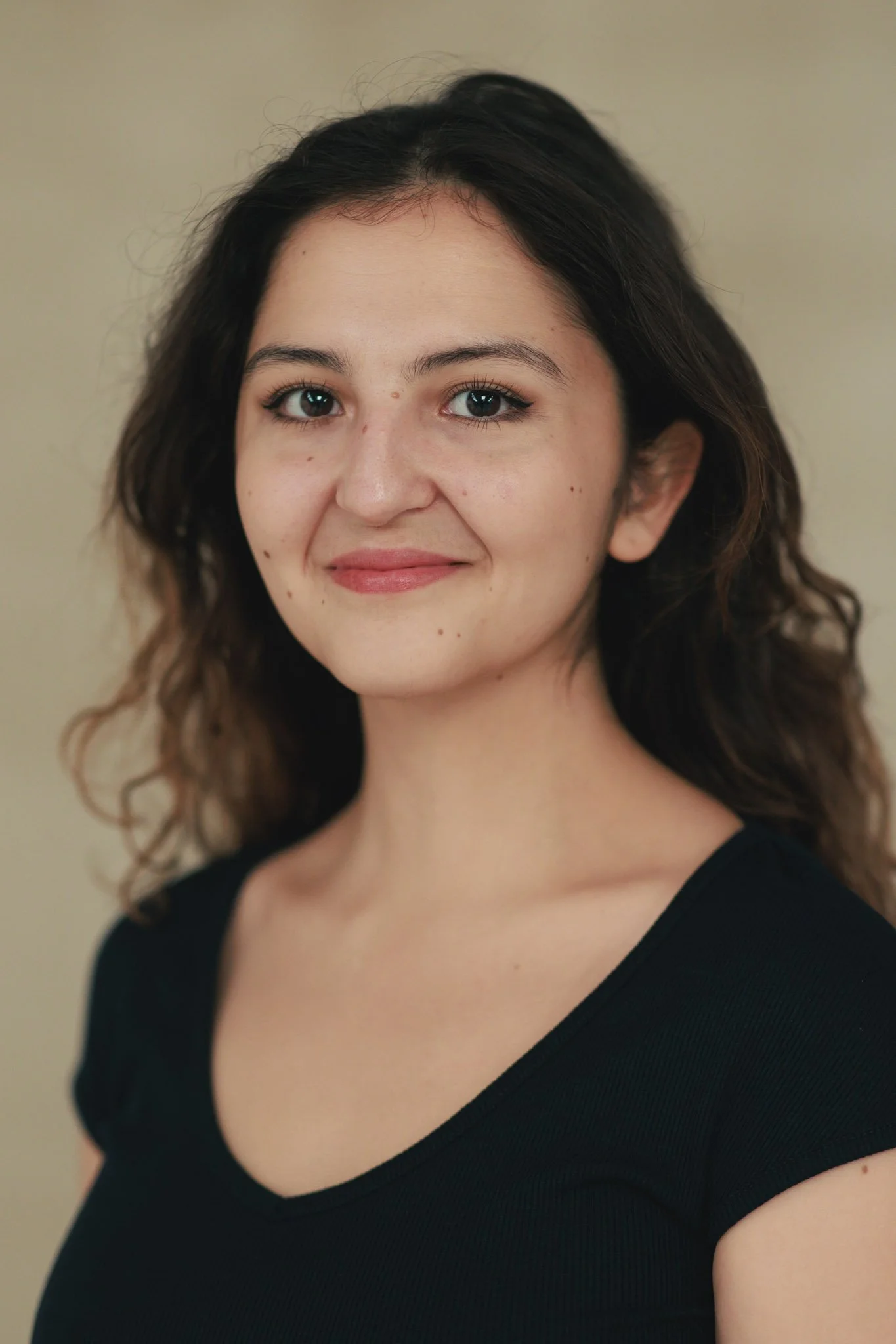 A woman with shoulder-length dark hair, light skin, and wearing a black top, smiling gently at the camera against a plain beige background.