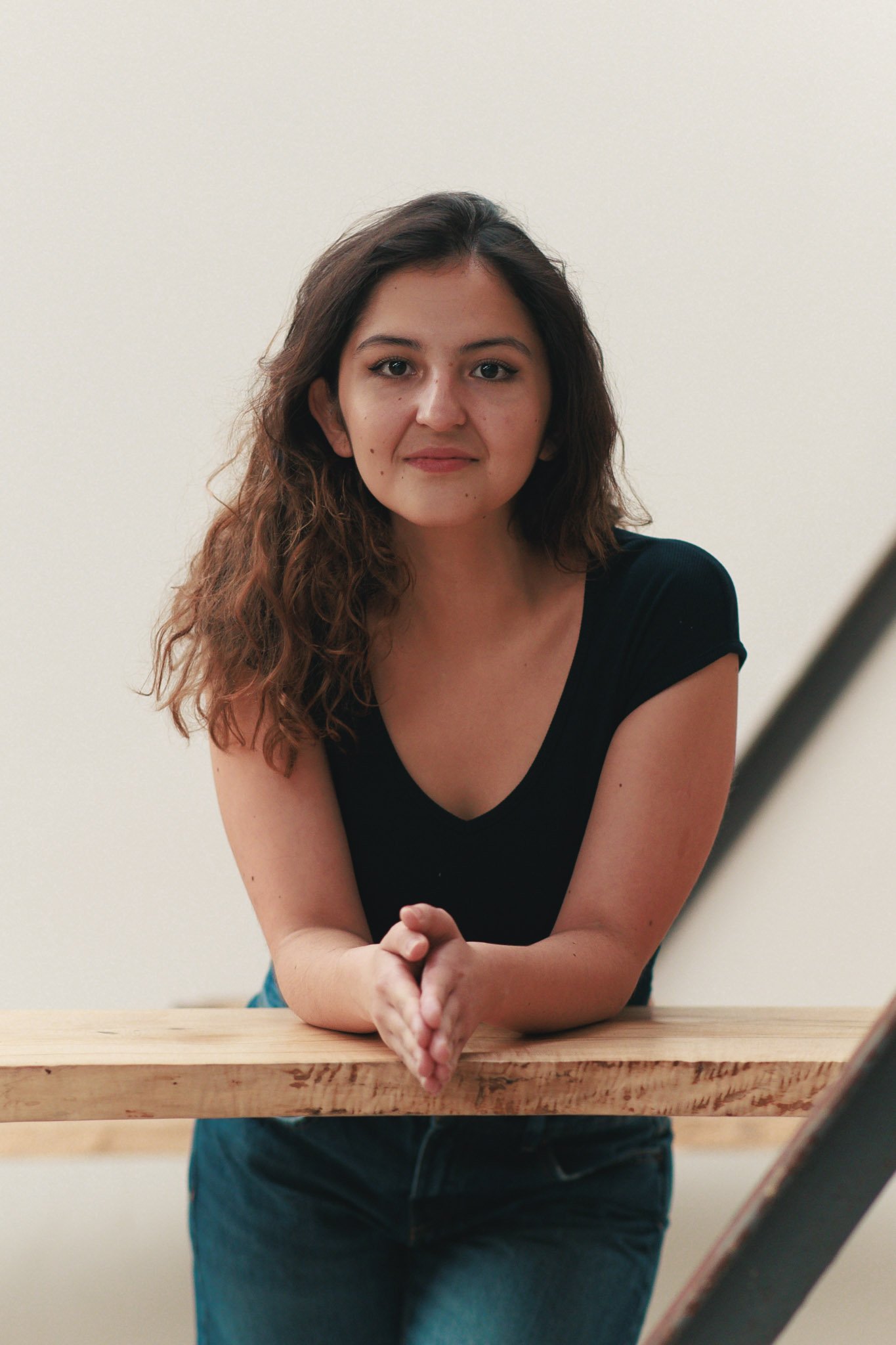 A woman with shoulder-length wavy brown hair, wearing a black shirt, leaning on a wooden barrier with her hands clasped, in front of a plain off-white wall.
