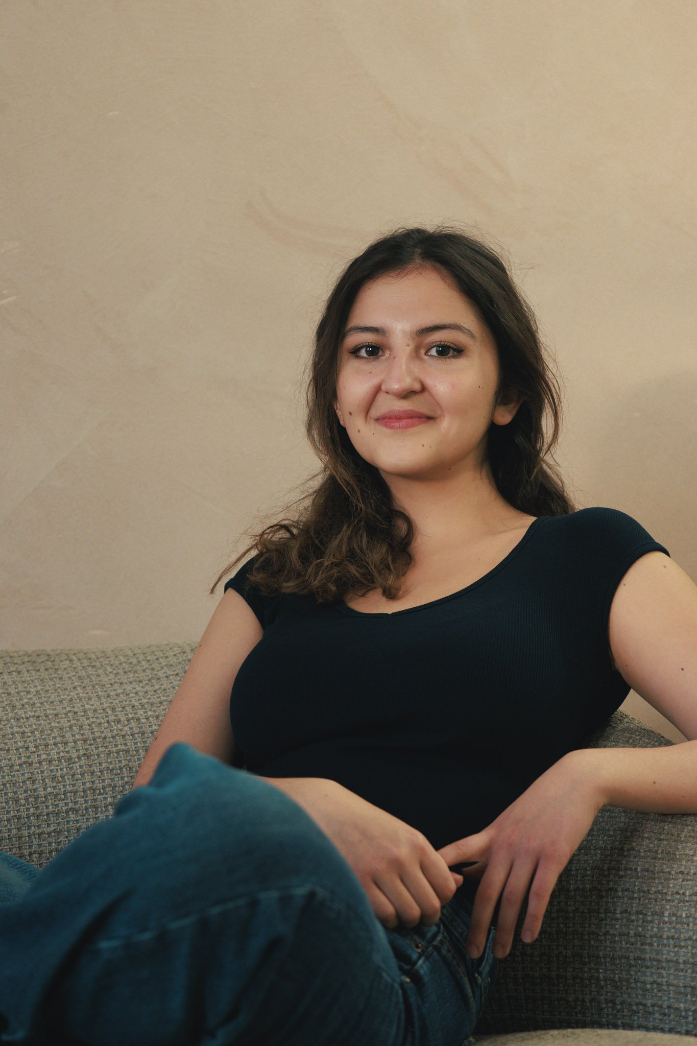 A young woman with shoulder-length wavy hair, wearing a black top and blue jeans, sitting on a beige couch with a neutral wall background, smiling slightly at the camera.