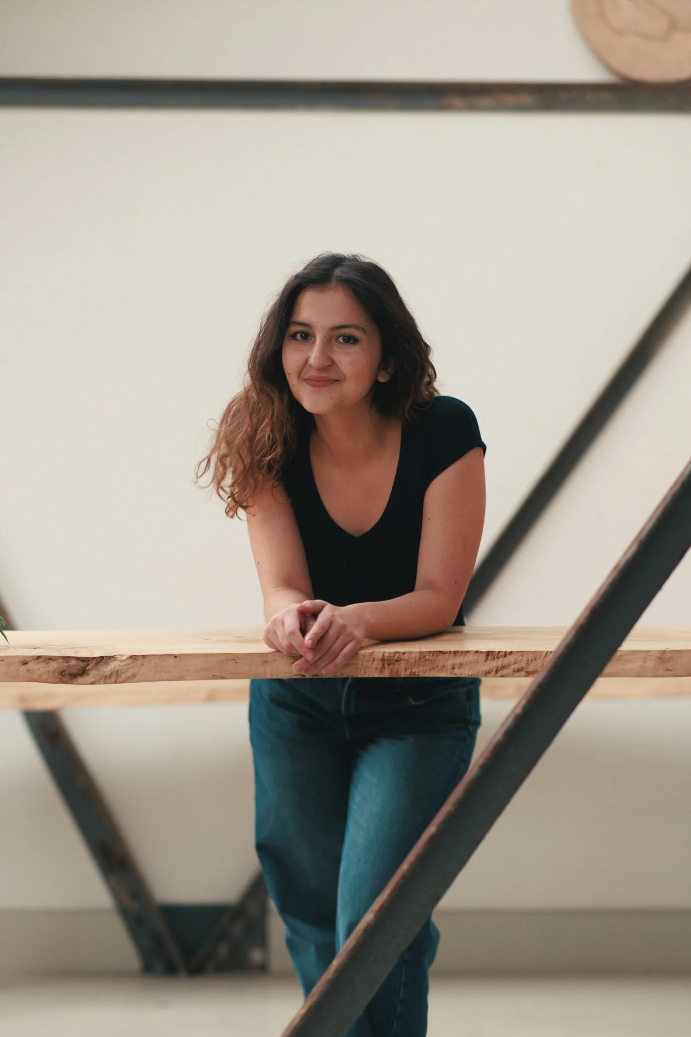 A young woman with curly brown hair, wearing a black T-shirt and blue jeans, leaning on a wooden surface with a neutral background.