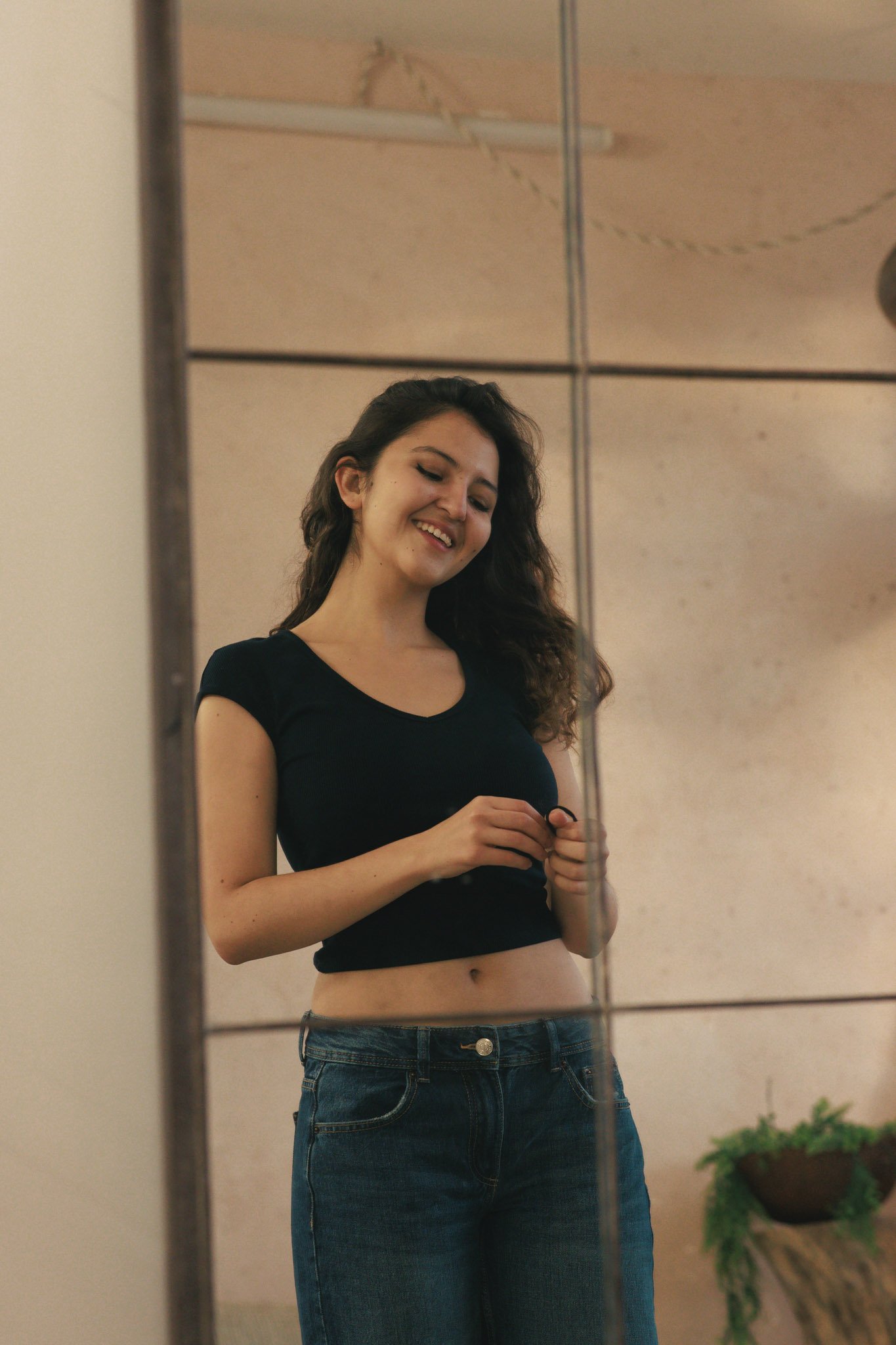 A woman smiles while taking a mirror selfie, holding a phone in her hands. She has long curly hair and is wearing a black crop top and blue jeans. The background includes a wooden ceiling and a plant in a brown container.