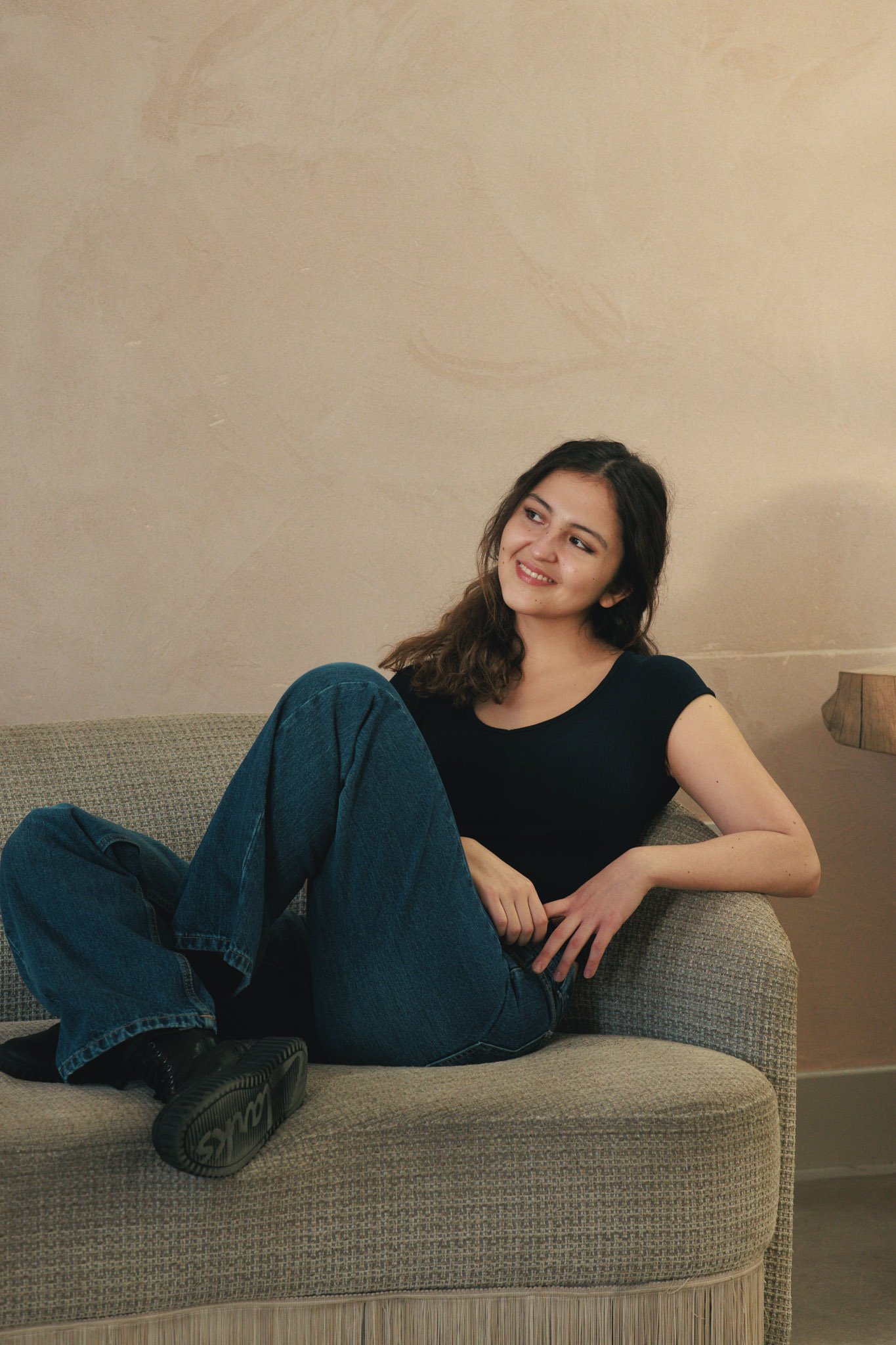 A young woman with wavy brown hair sits comfortably on a sofa, wearing a black t-shirt, blue jeans, and black shoes, smiling and looking to her left in a warmly lit room with a beige wall.