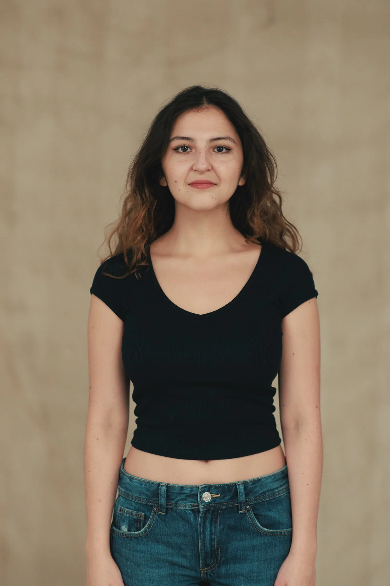 A young woman with shoulder-length brown, wavy hair, wearing a black short-sleeved crop top and blue jeans, standing against a neutral background.