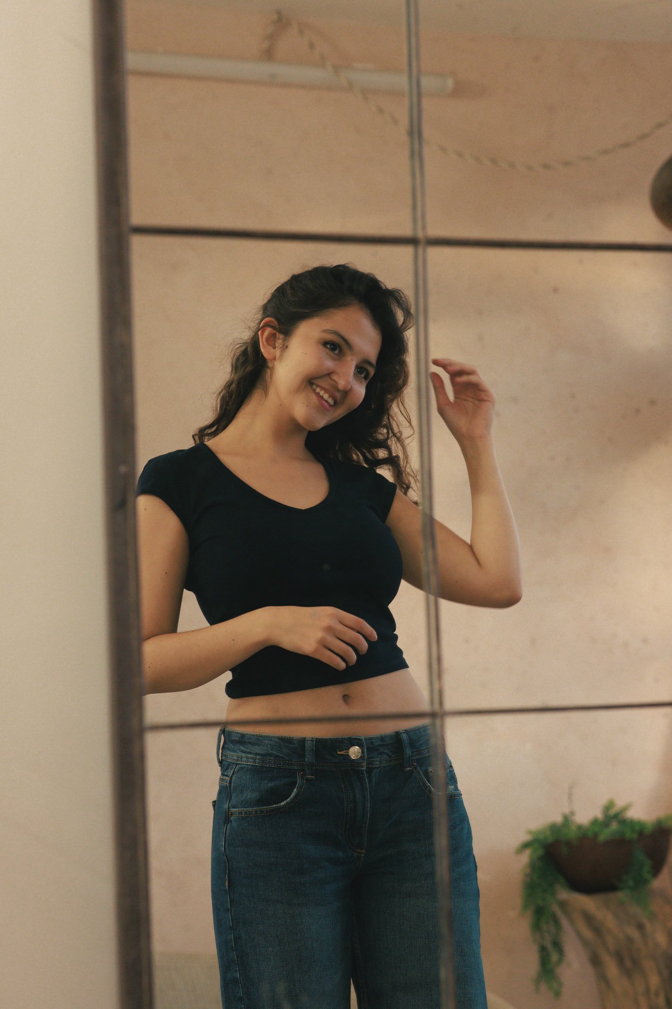 A young woman with curly brown hair and fair skin taking a mirror selfie. She is smiling and touching her hair, wearing a black crop top and blue jeans. There is a potted plant visible in the background.