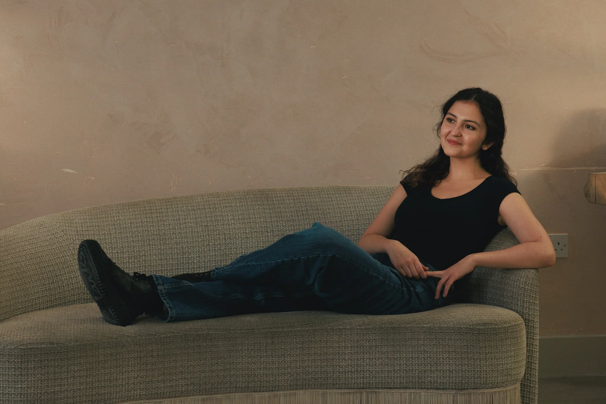 A woman with dark wavy hair sitting on a beige couch, wearing a black T-shirt, blue jeans, and black boots, smiling softly. The wall behind her is beige with some texture, and there is a lamp on the right side of the image.