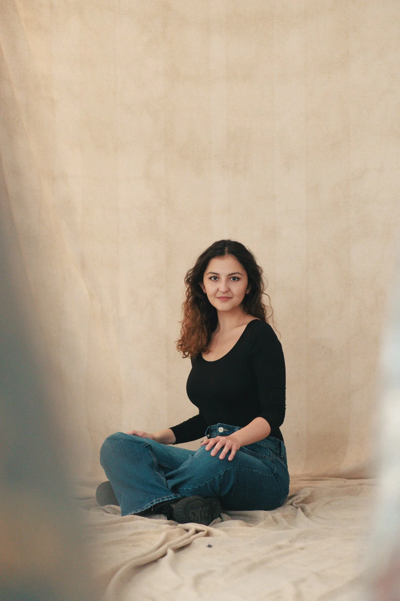 A young woman with long, curly brown hair sits cross-legged on a beige blanket in front of a plain beige backdrop. She is wearing a black long-sleeve top and blue jeans, and is looking directly at the camera with a neutral expression.