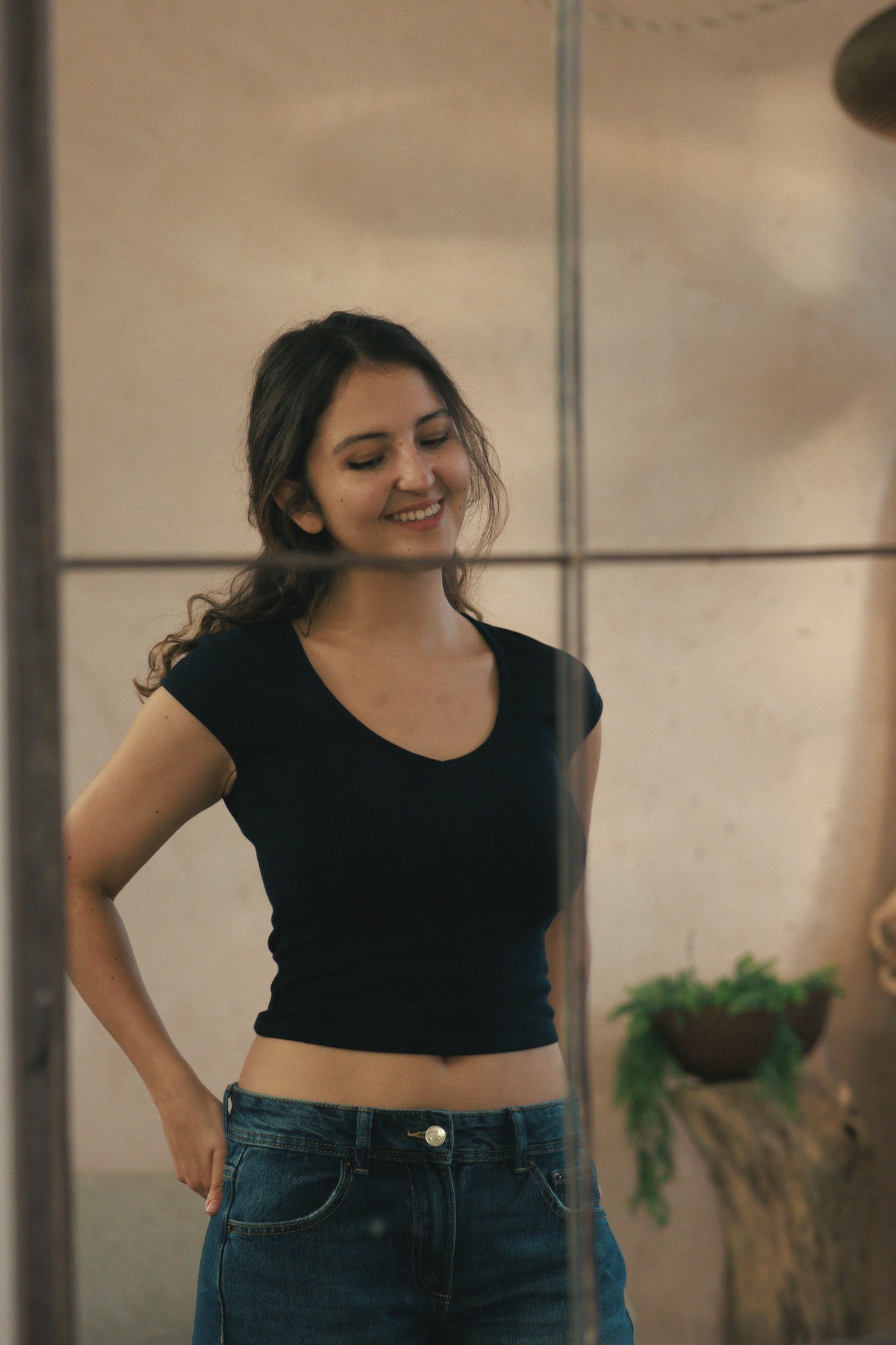 Young woman with curly brown hair smiling while looking at her reflection in a mirror, wearing a black short-sleeve shirt and blue jeans.
