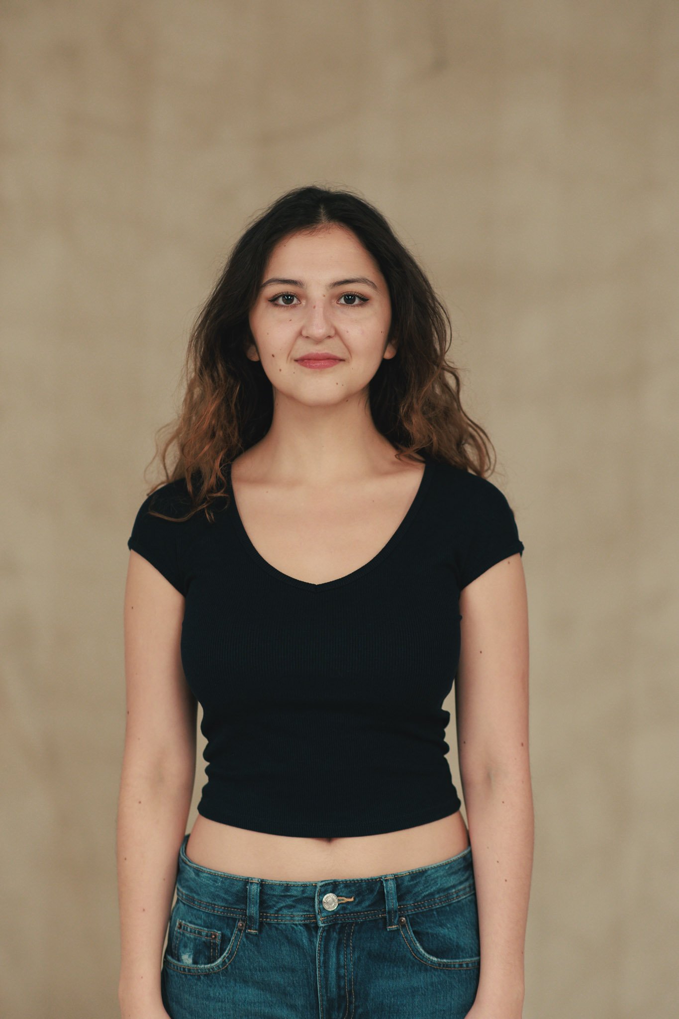 A young woman with wavy brown hair wearing a black crop top and blue jeans, standing against a beige background.