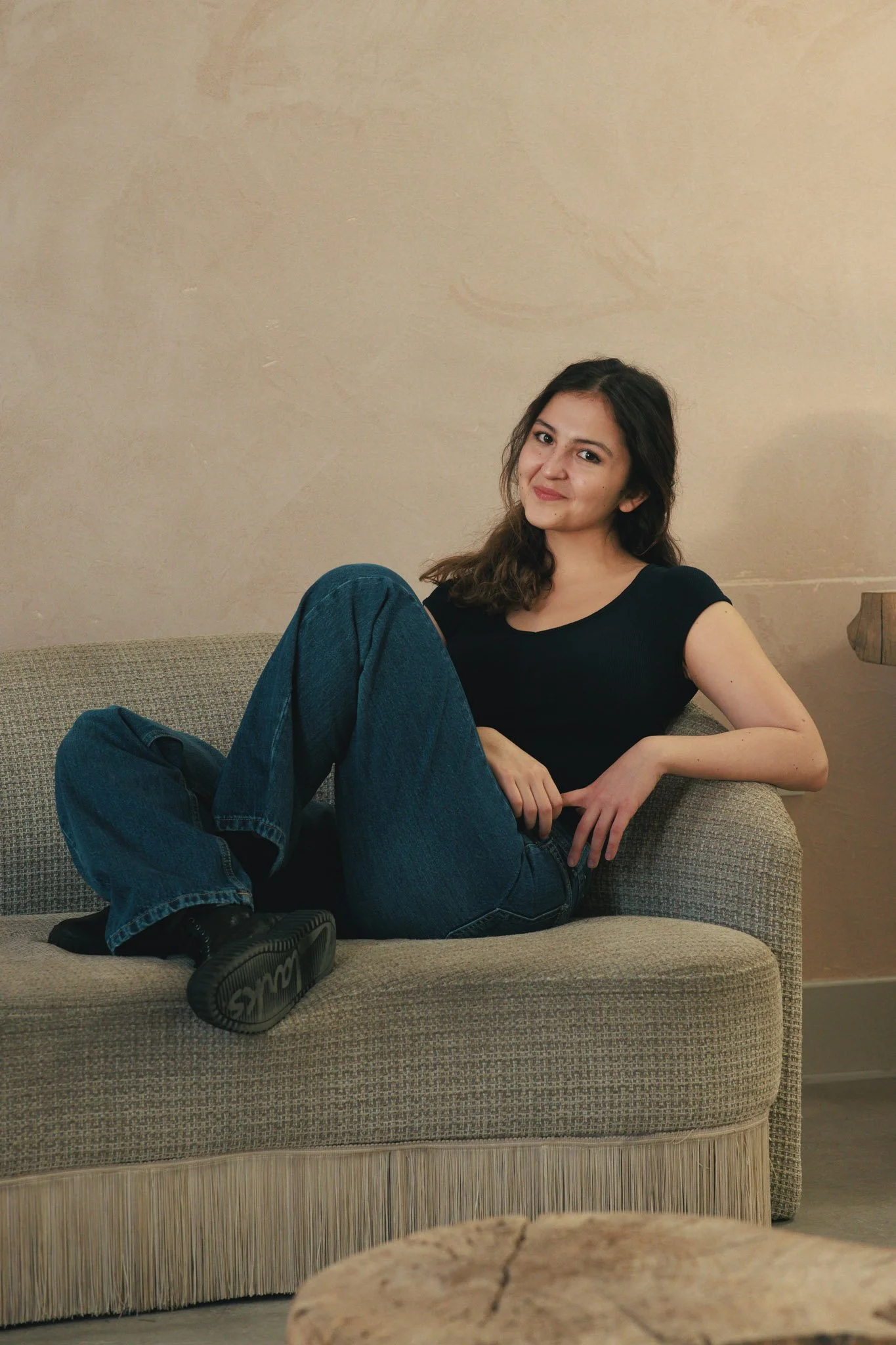 A young woman with dark hair, wearing a black t-shirt, jeans, and black shoes, is sitting relaxed on a beige sofa with a textured fabric and fringe at the bottom. She is leaning back, with one arm resting on the sofa's armrest and the other hand on h