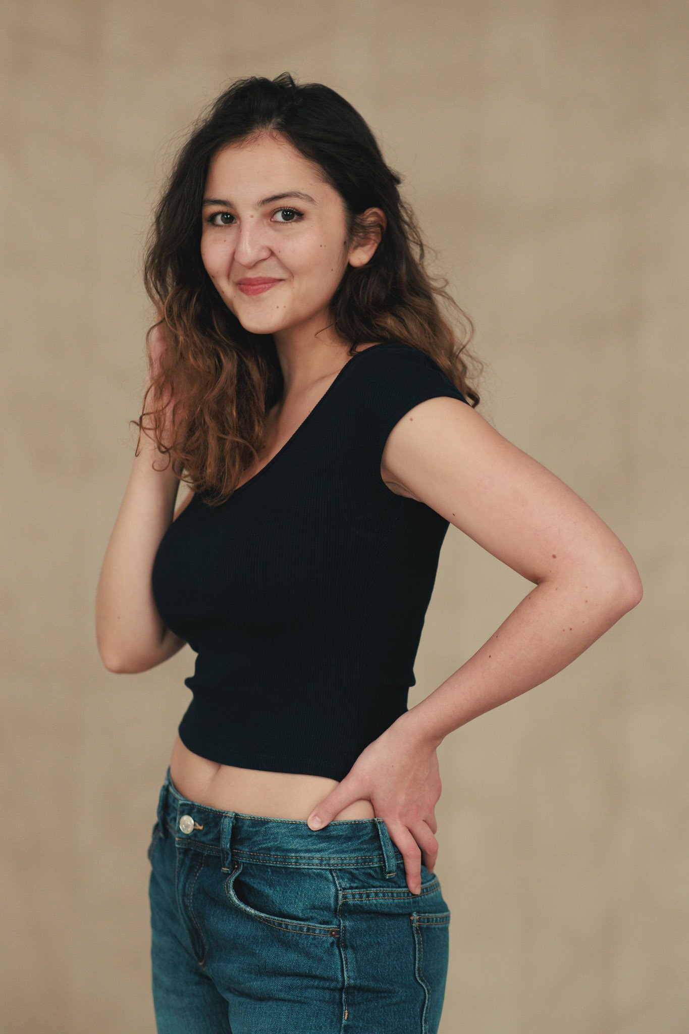 Young woman with long, wavy brown hair smiling, wearing a black sleeveless top and blue jeans, standing with one hand on her hip against a beige background.