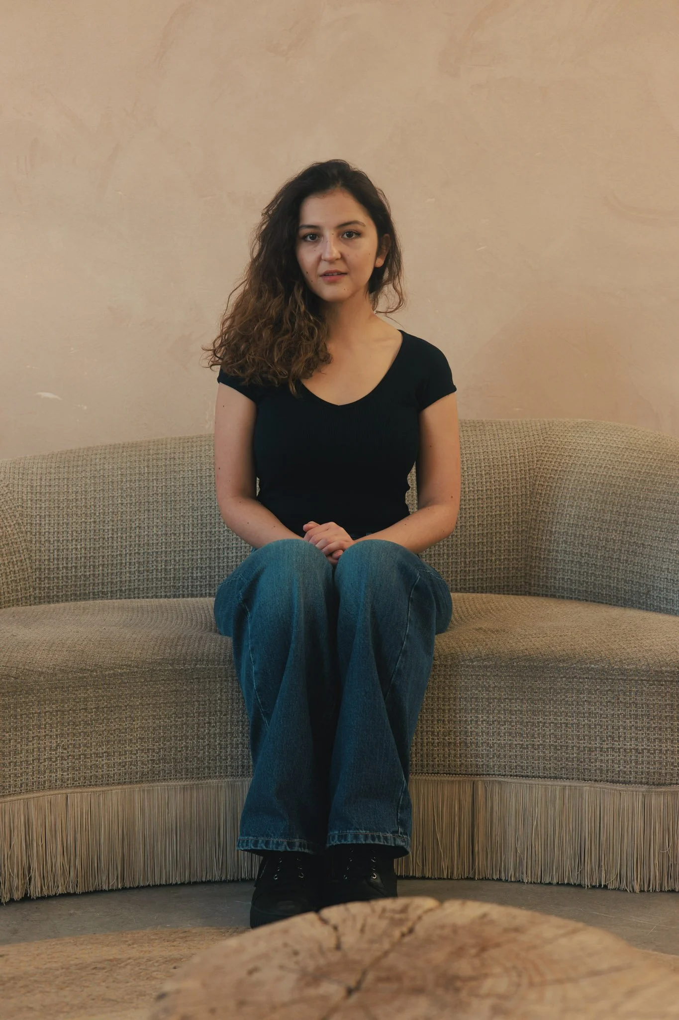 A woman with curly hair wearing a black t-shirt and blue jeans, sitting on a beige sofa against a plain wall.