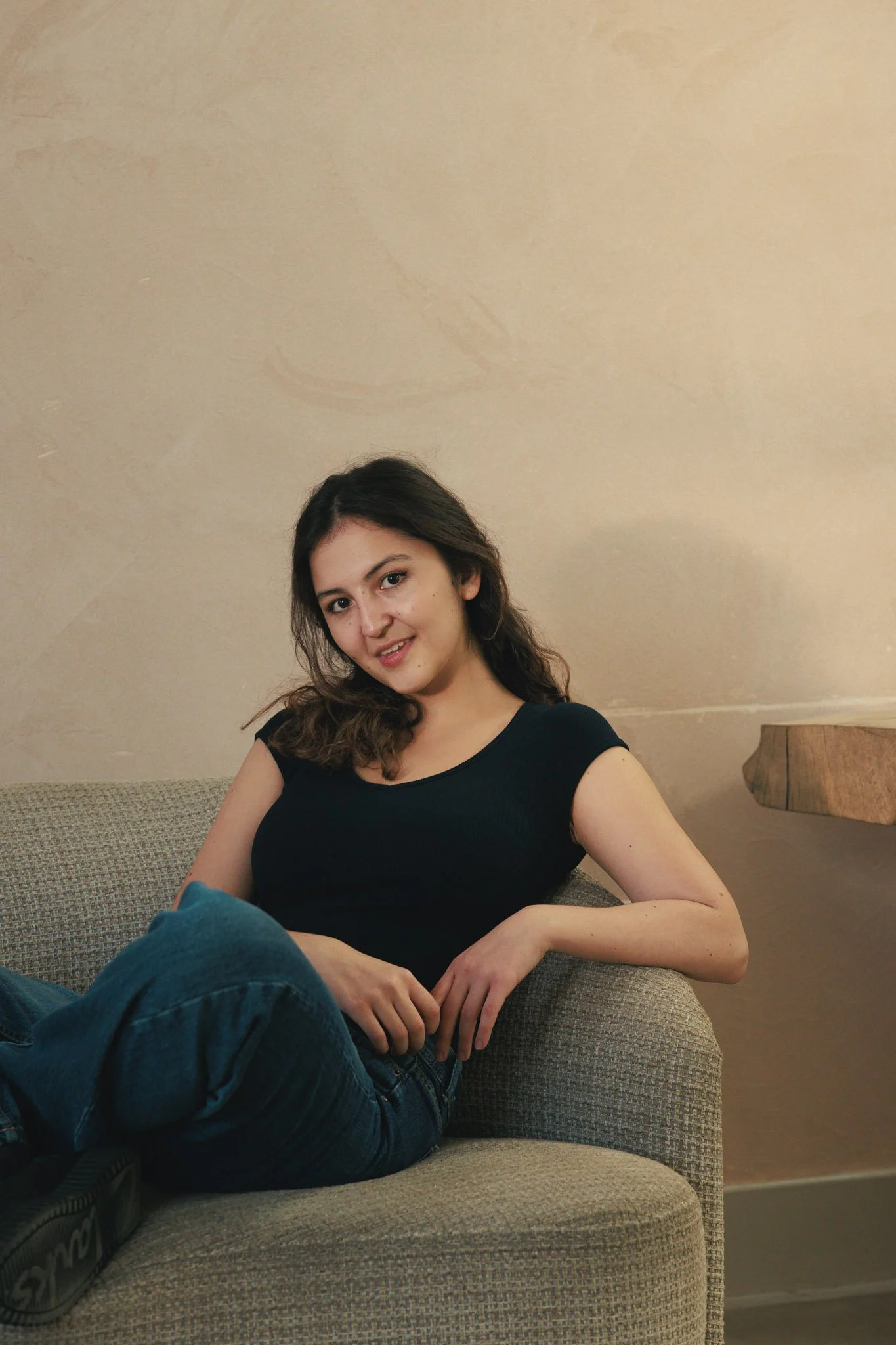 Young woman with curly hair in a black t-shirt and blue jeans sitting relaxed on a beige sofa in a room with plain wall and wooden detail.