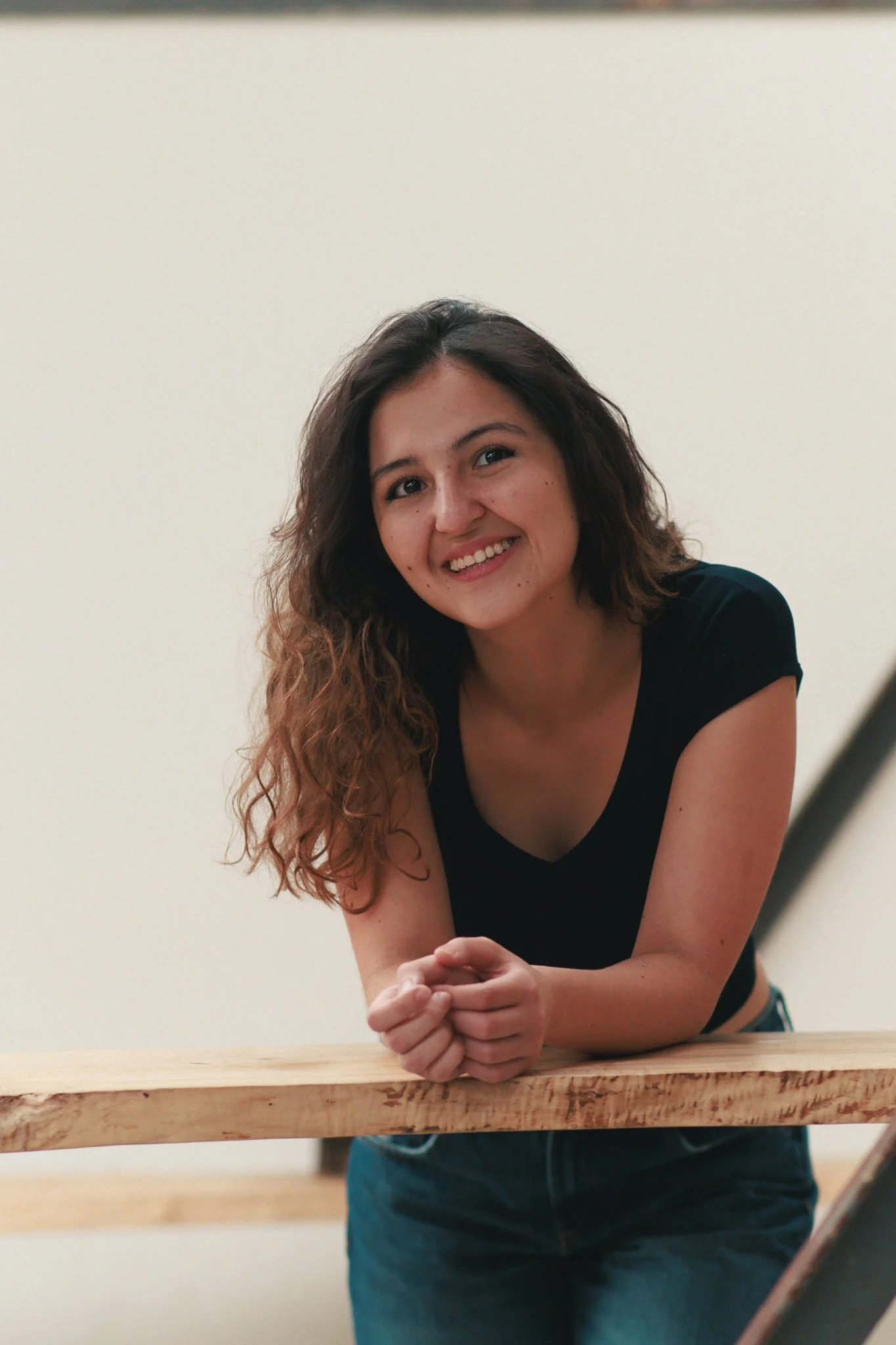 A woman with wavy brown hair smiling and leaning on a wooden railing with a plain white wall background.