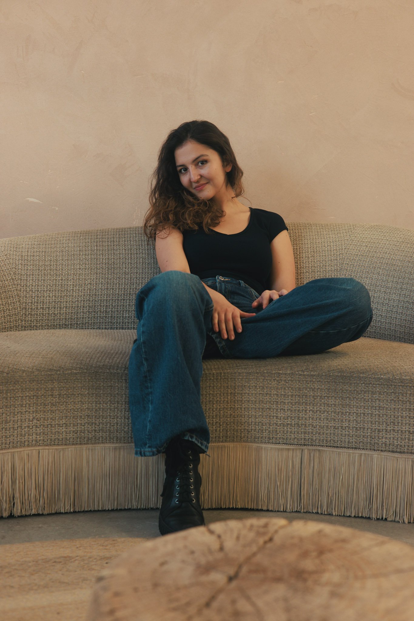 A young woman with curly hair, sitting cross-legged on a beige couch, wearing a black shirt, jeans, and black boots, with a smile, in a room with a beige wall.