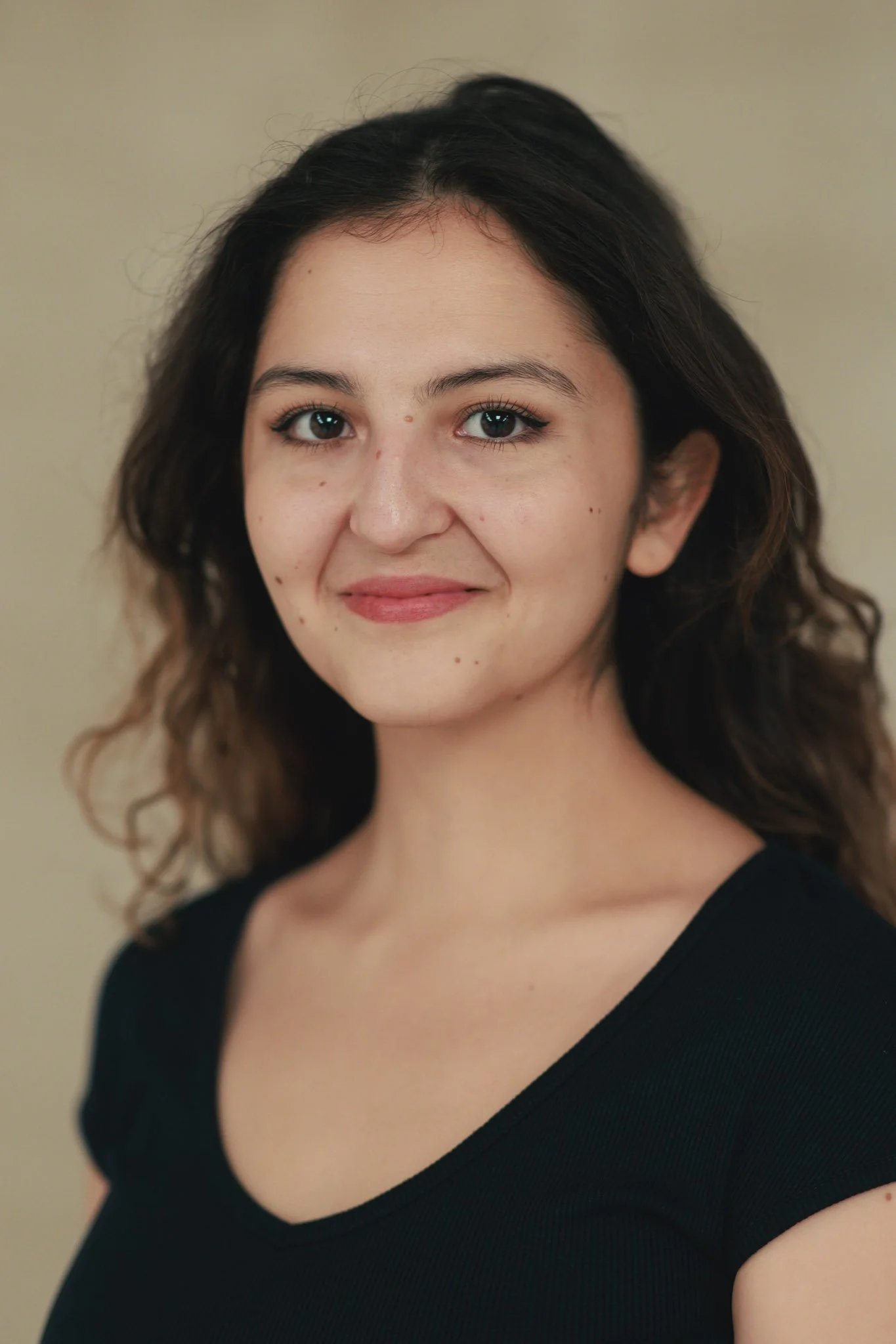 Close-up portrait of a young woman with brown curly hair, wearing a black top, smiling softly against a neutral background.