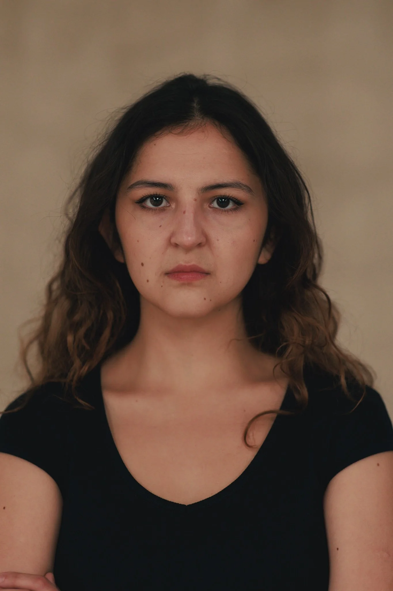 Portrait of a young woman with wavy brown hair, wearing a black shirt, looking directly at the camera against a neutral background.