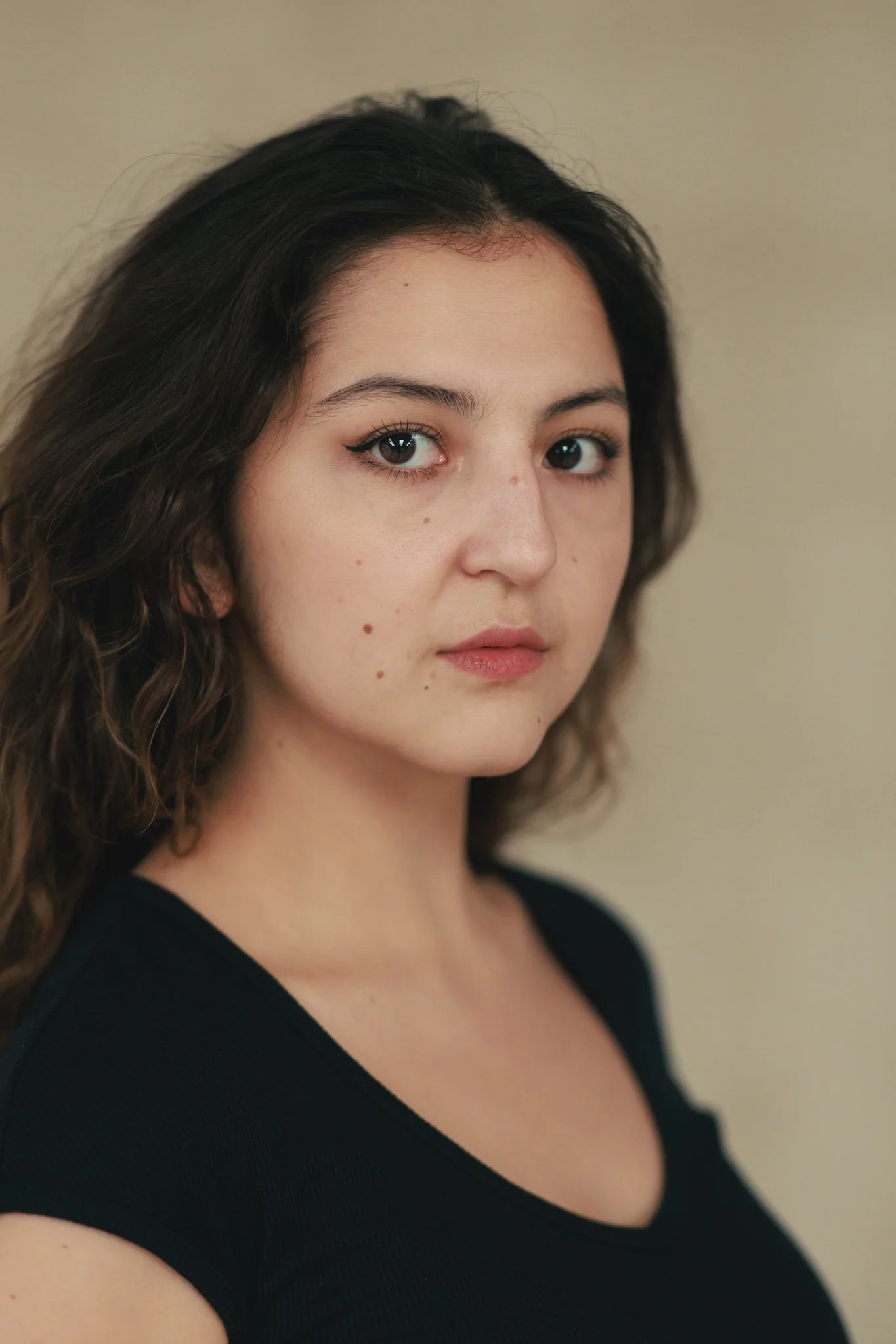 A portrait of a young woman with wavy dark hair and light skin, wearing a black top, standing against a neutral background.