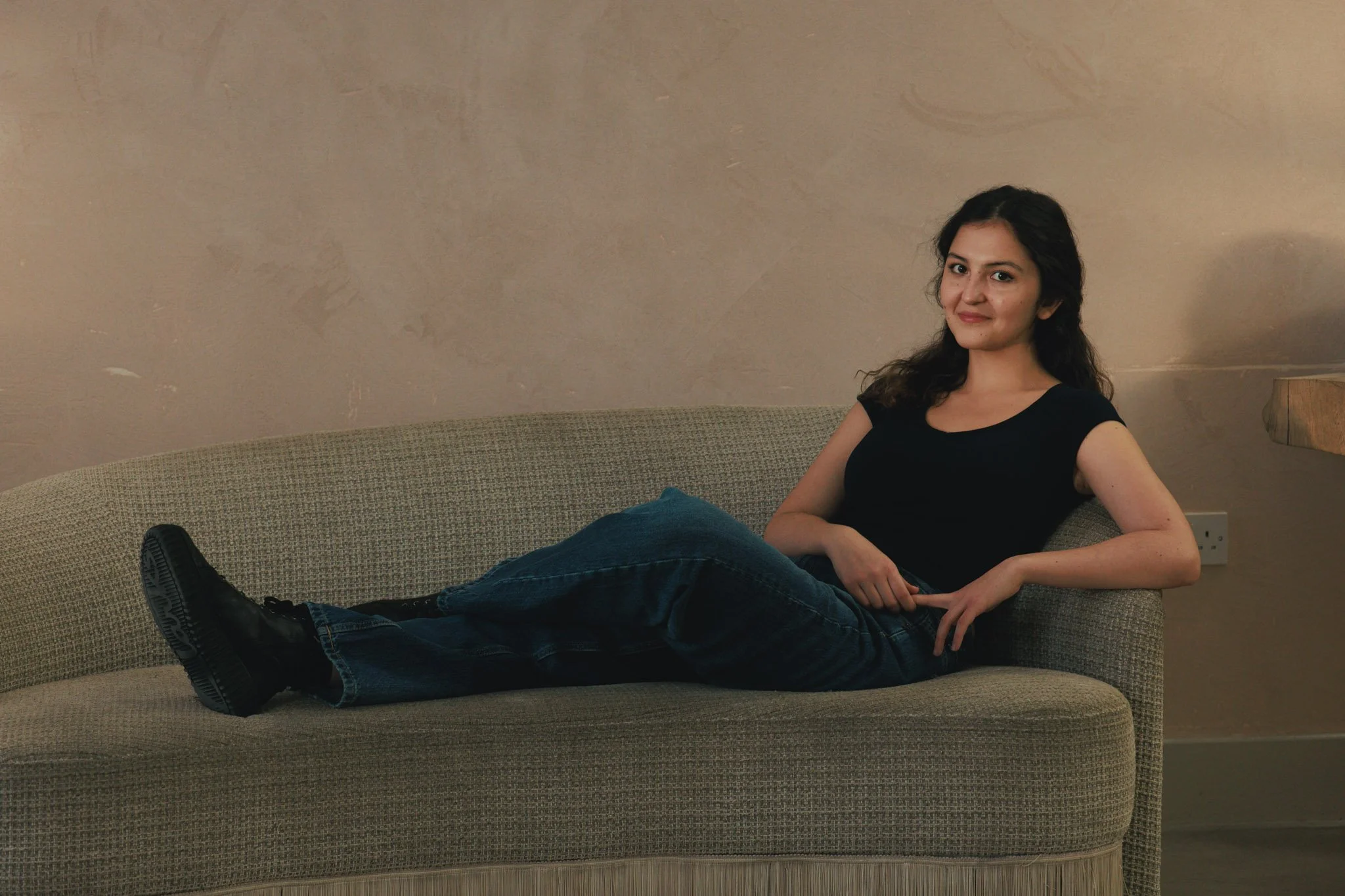 A woman with dark hair wearing a black t-shirt and dark jeans, lying on a beige sofa with one leg extended and the other bent, in a room with a plain wall and a side table.