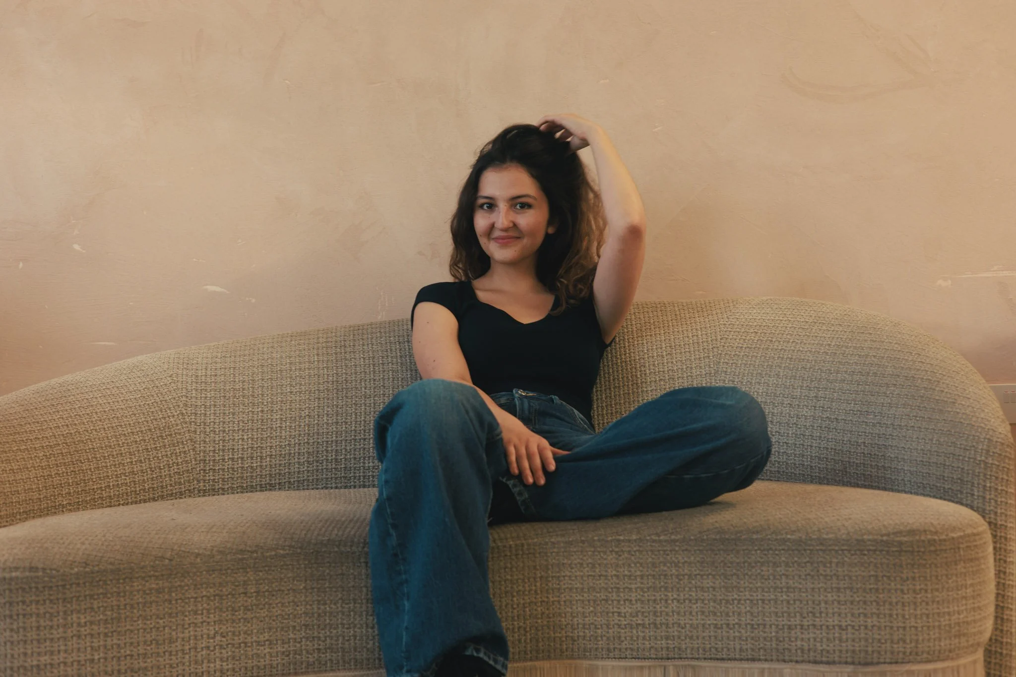 A woman with curly brown hair sitting on a beige couch, wearing a black t-shirt and blue jeans, smiling and touching her hair with her right hand, against a light-colored wall.