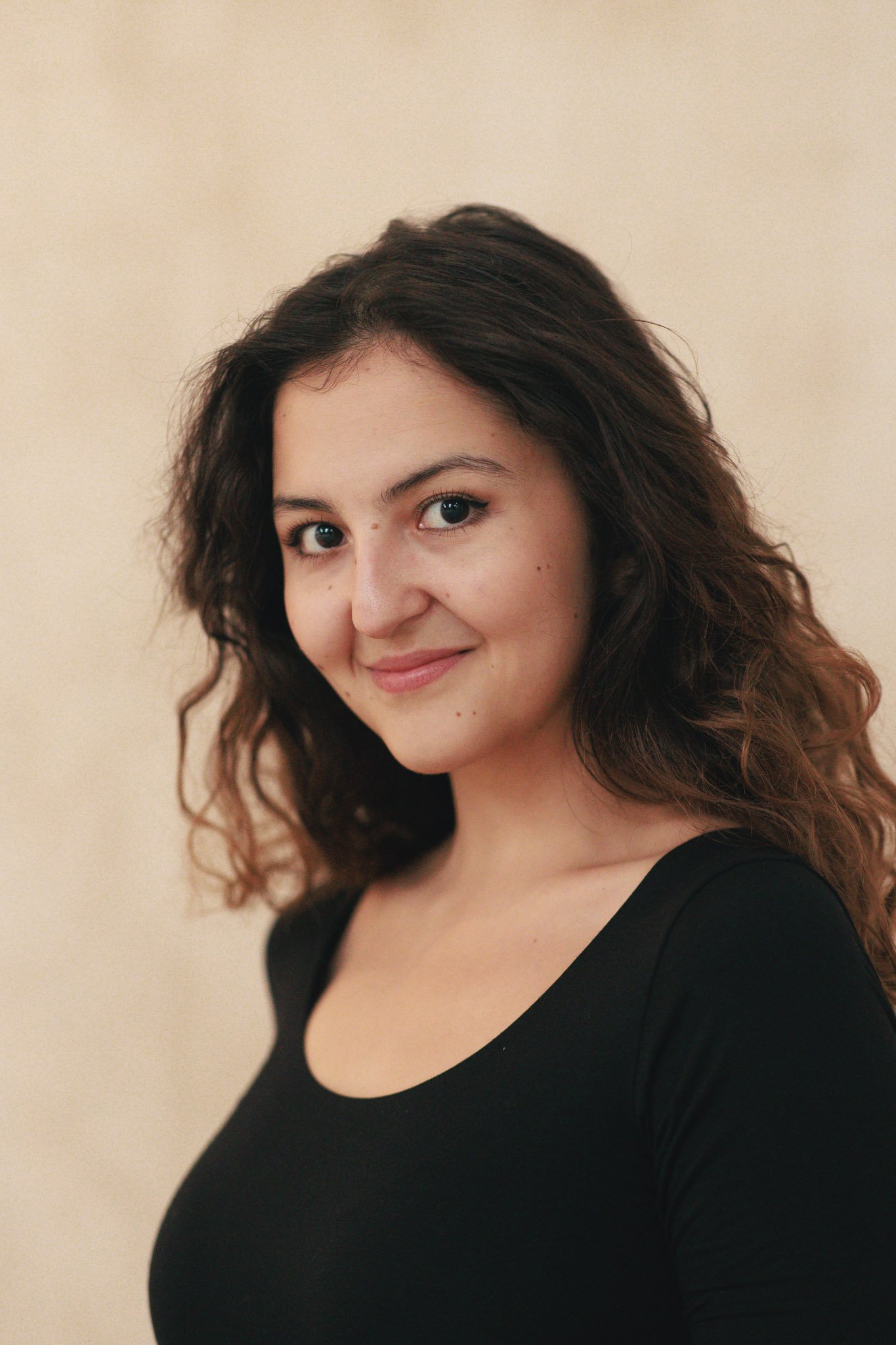 A young woman with wavy dark hair and blue eyes, wearing a black top, smiling at the camera against a beige background.