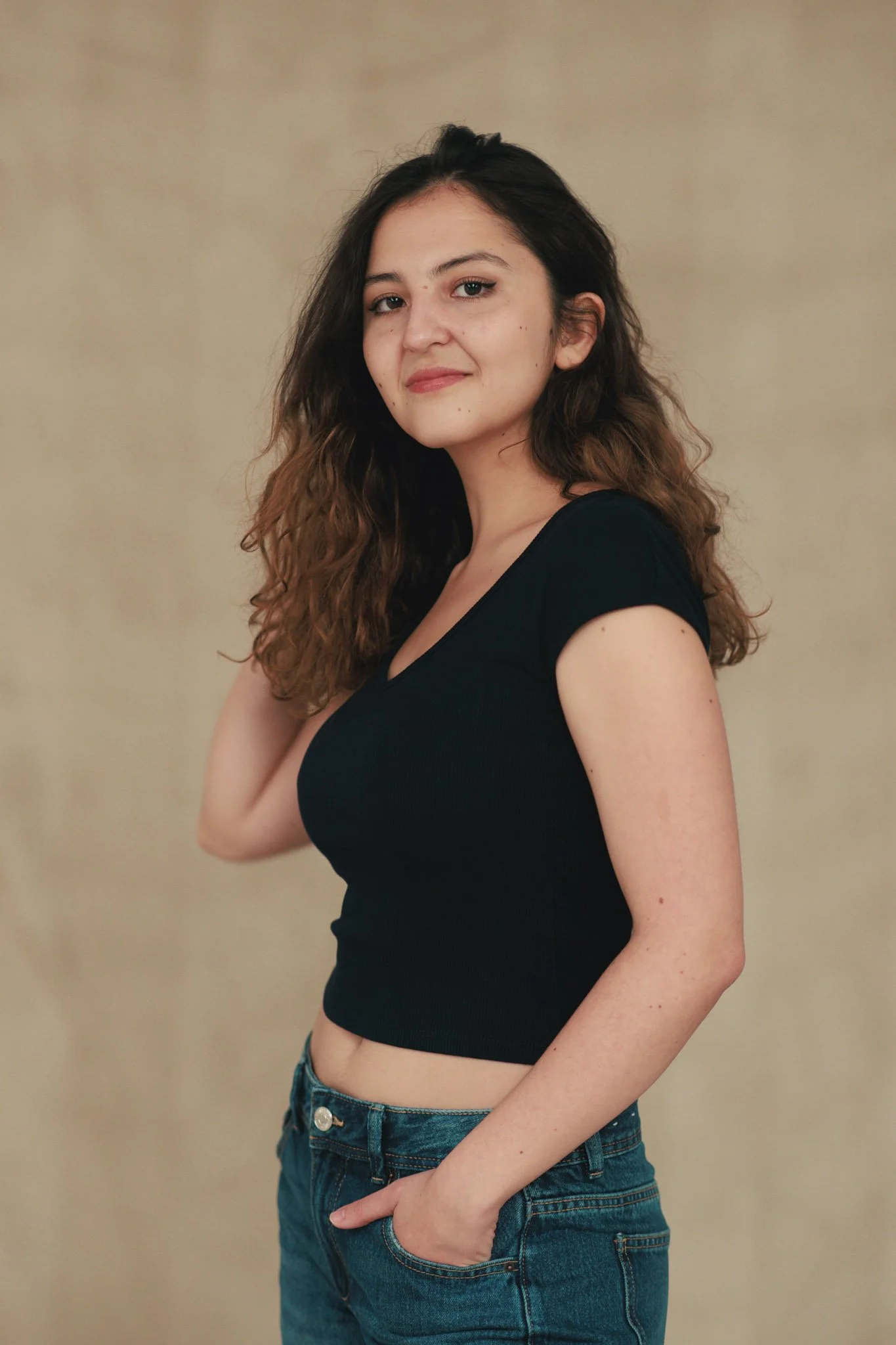 A young woman with long, wavy brown hair, wearing a black cropped t-shirt and blue jeans, standing with her left hand in her pocket against a beige background, looking at the camera with a slight smile.