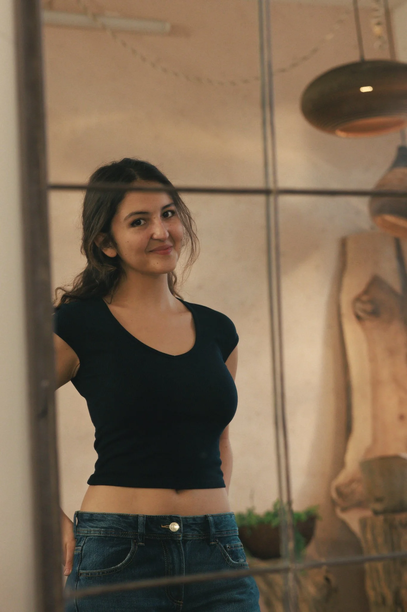 A young woman with dark hair wearing a black crop top and blue jeans, standing indoors, looking into a mirror with a slight smile.