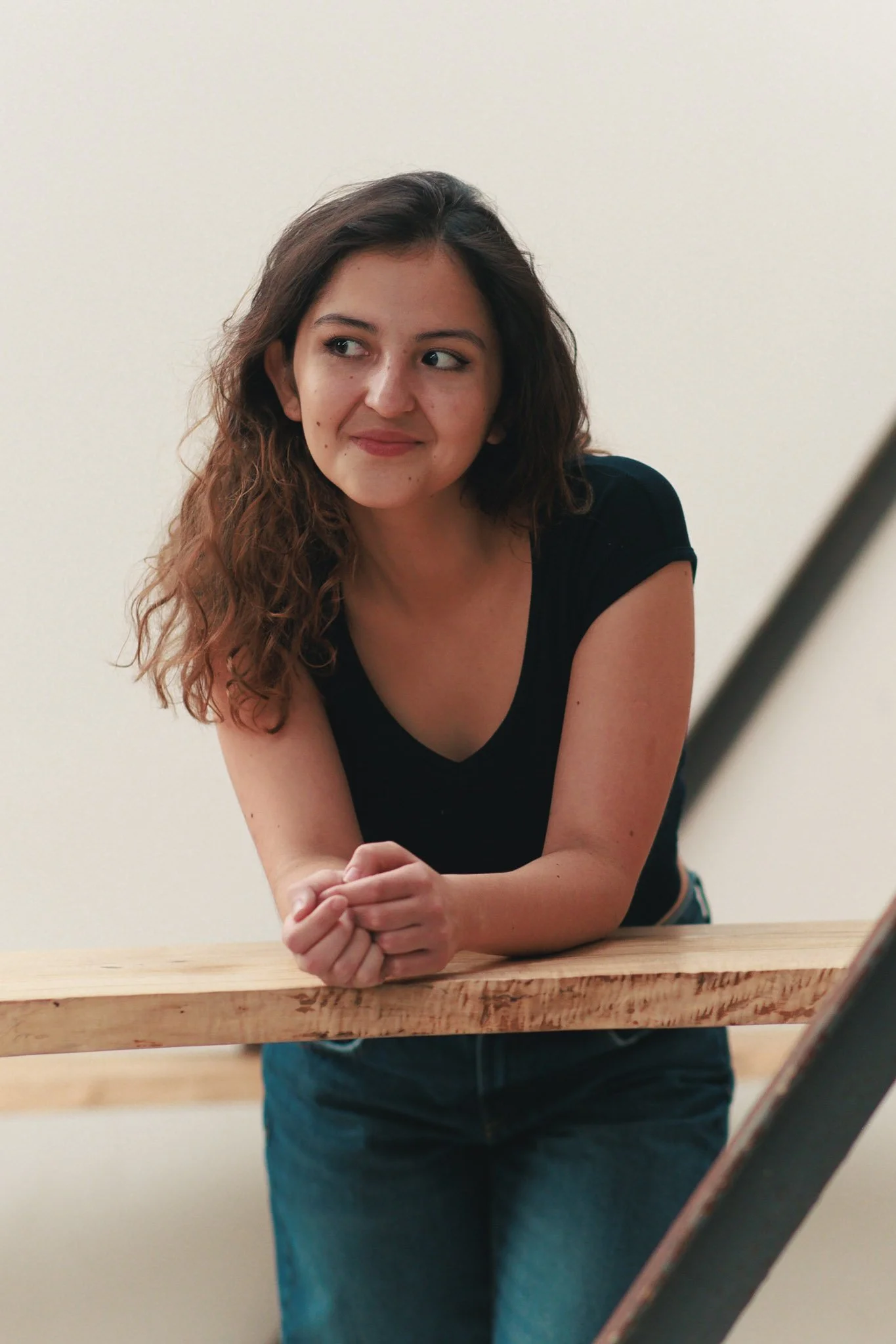 A woman with curly brown hair wearing a black t-shirt leaning on a wooden railing, looking to her left with a slight smile.