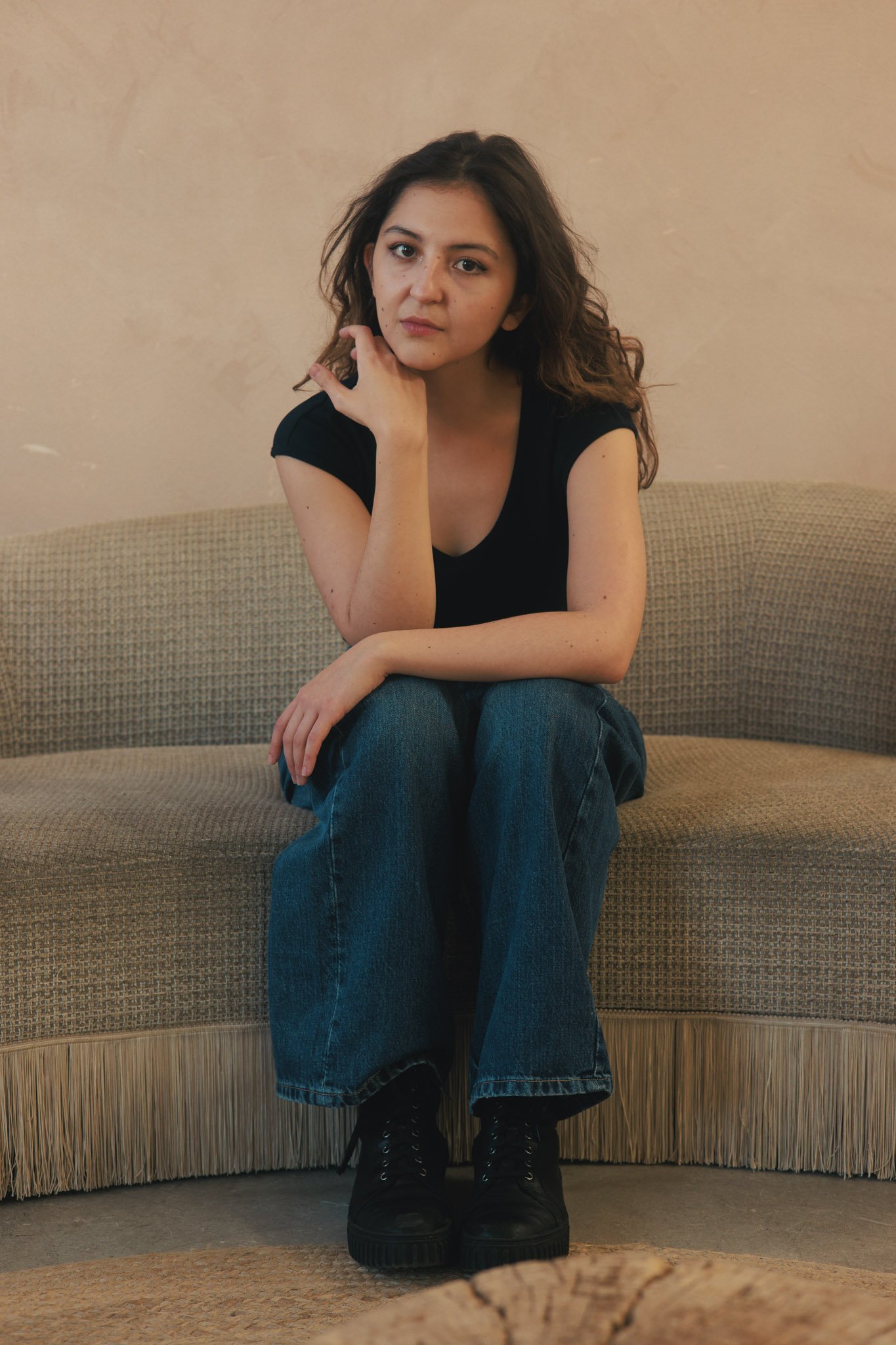A young woman with wavy brown hair sitting on a beige textured couch in a room with a plain cream wall, wearing a black t-shirt, blue jeans, and black boots. She is resting her chin on her left hand and looking directly at the camera.
