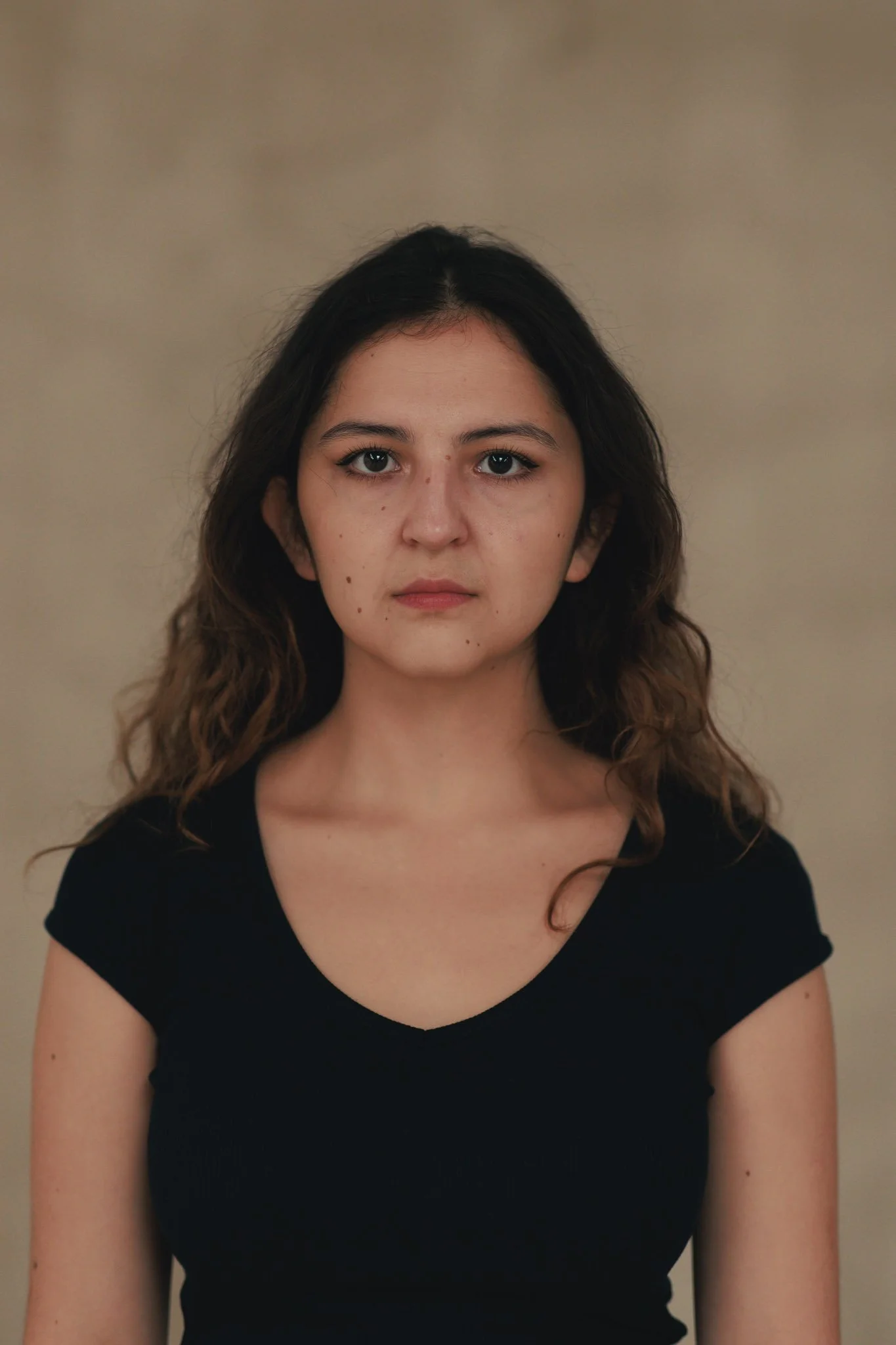 A young woman with wavy brown hair, wearing a black shirt, looking directly at the camera with a neutral expression against a plain beige background.