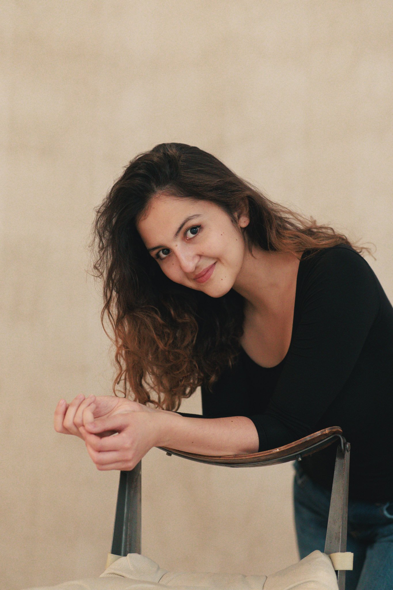 A young woman with long curly brown hair wearing a black top, leaning on the back of a chair with a neutral beige wall in the background.