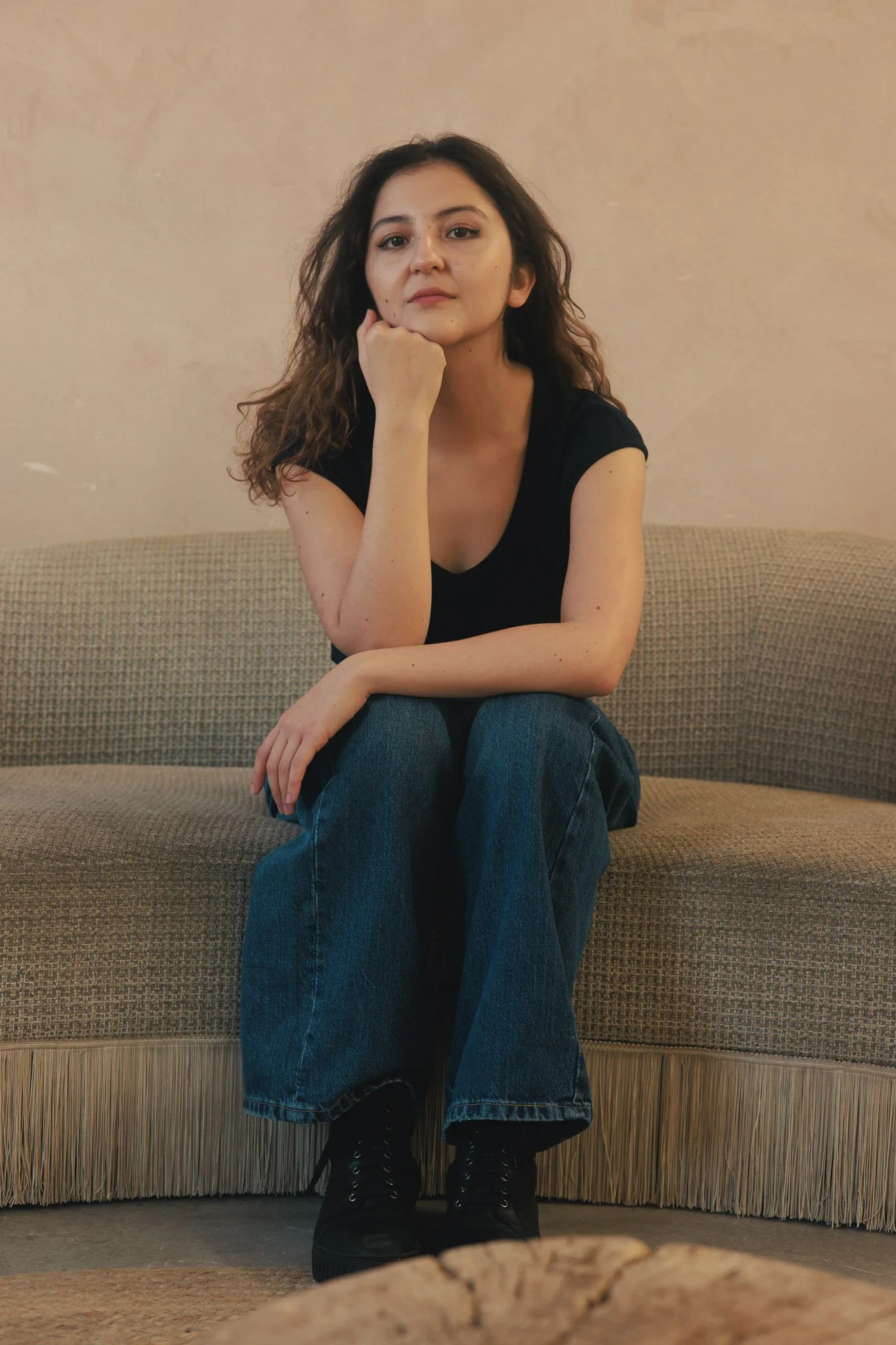 A young woman with wavy brown hair sitting on a beige sofa with fringe at the bottom, wearing a black T-shirt, blue jeans, and black boots, resting her chin on her hand and looking at the camera.