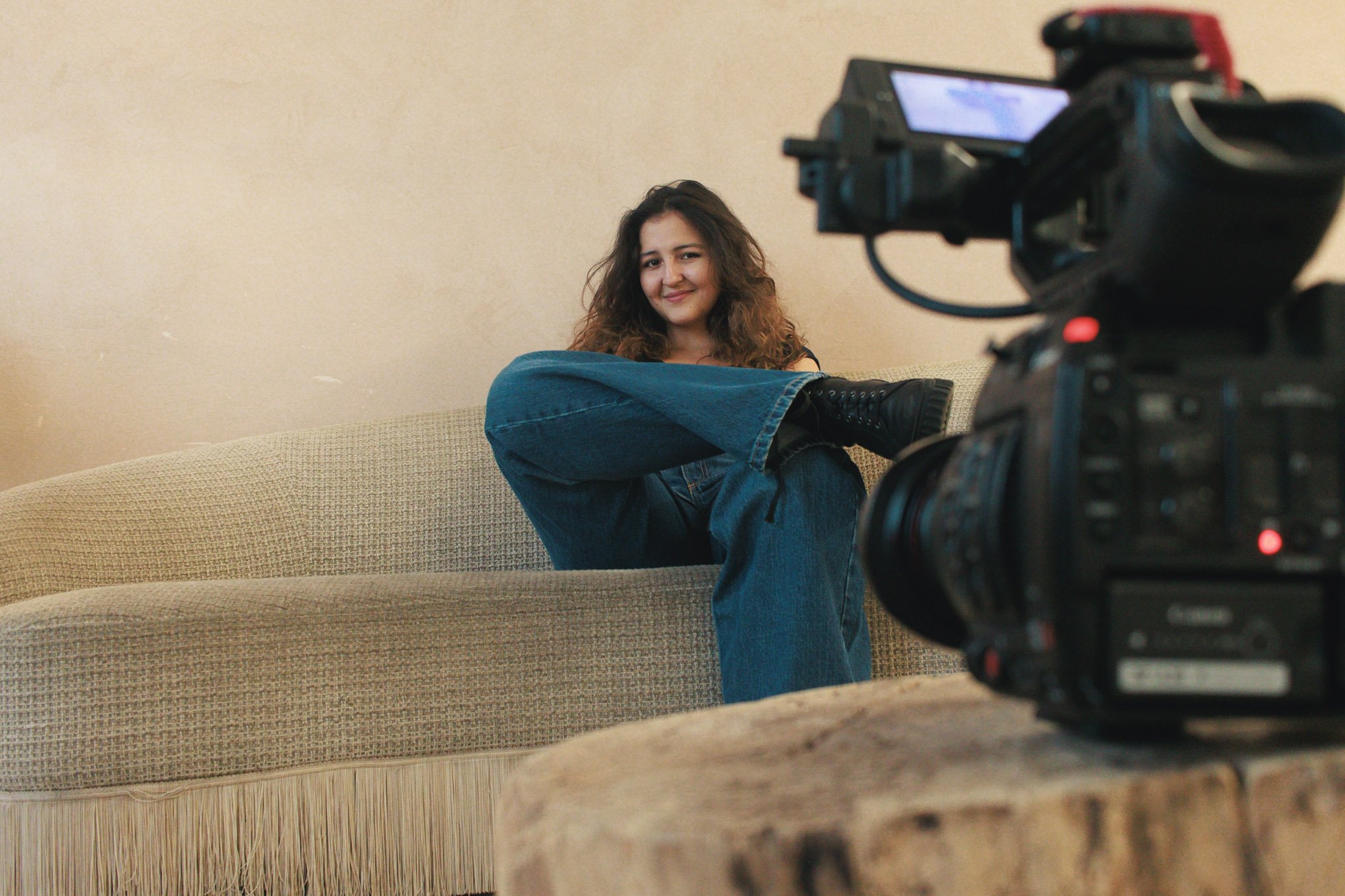A young woman with curly hair sitting on a beige sofa, smiling, with a video camera pointed at her.