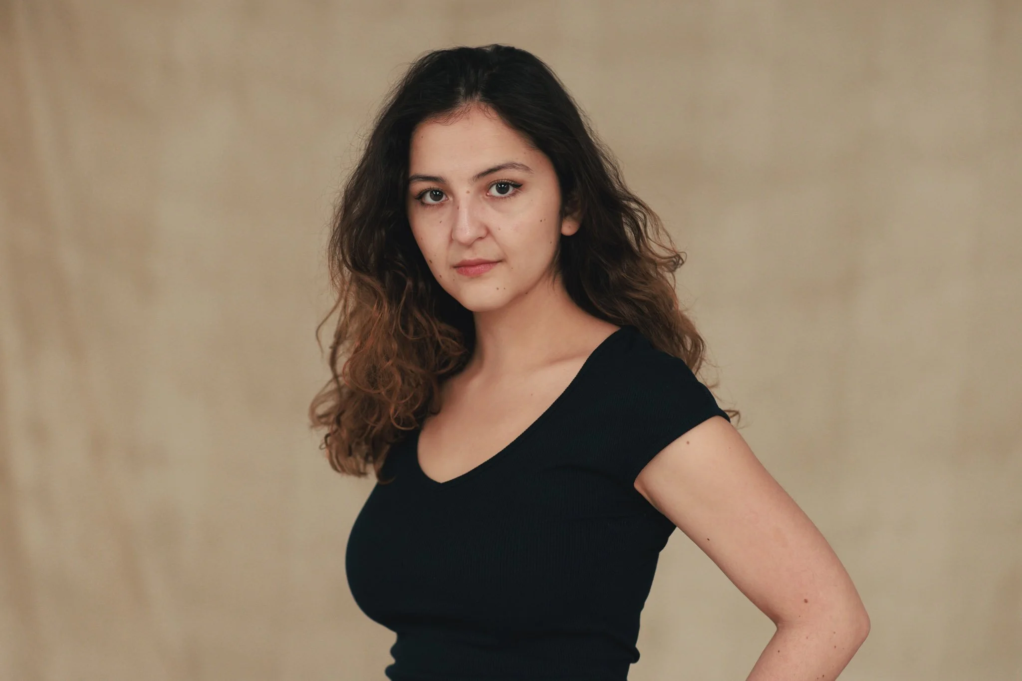 A young woman with long, curly brown hair and fair skin wearing a black top, standing against a beige background, looking directly at the camera with a serious expression.