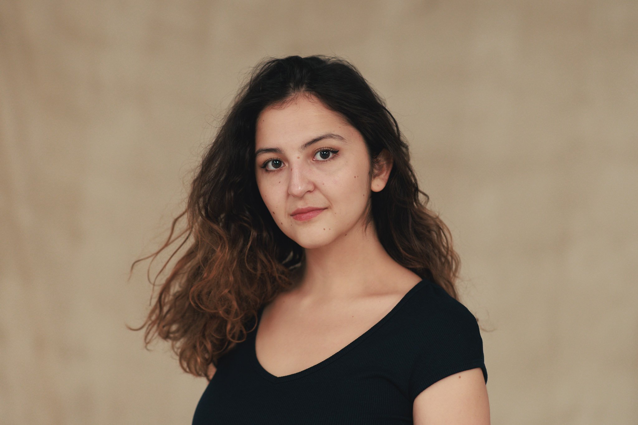 A young woman with wavy brown hair and light skin wearing a black top, posing in front of a beige background.