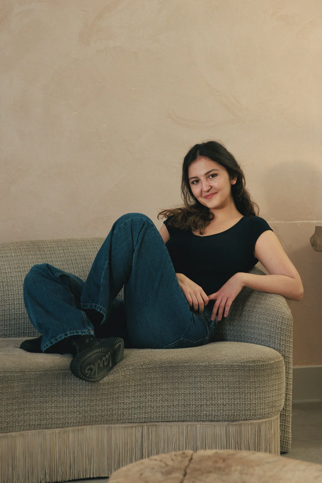 A young woman with wavy brown hair and light skin is sitting on a beige sofa with a textured fabric, wearing a black t-shirt, loose blue jeans, and black shoes. She is smiling at the camera with her right foot resting on the sofa and her left arm pro