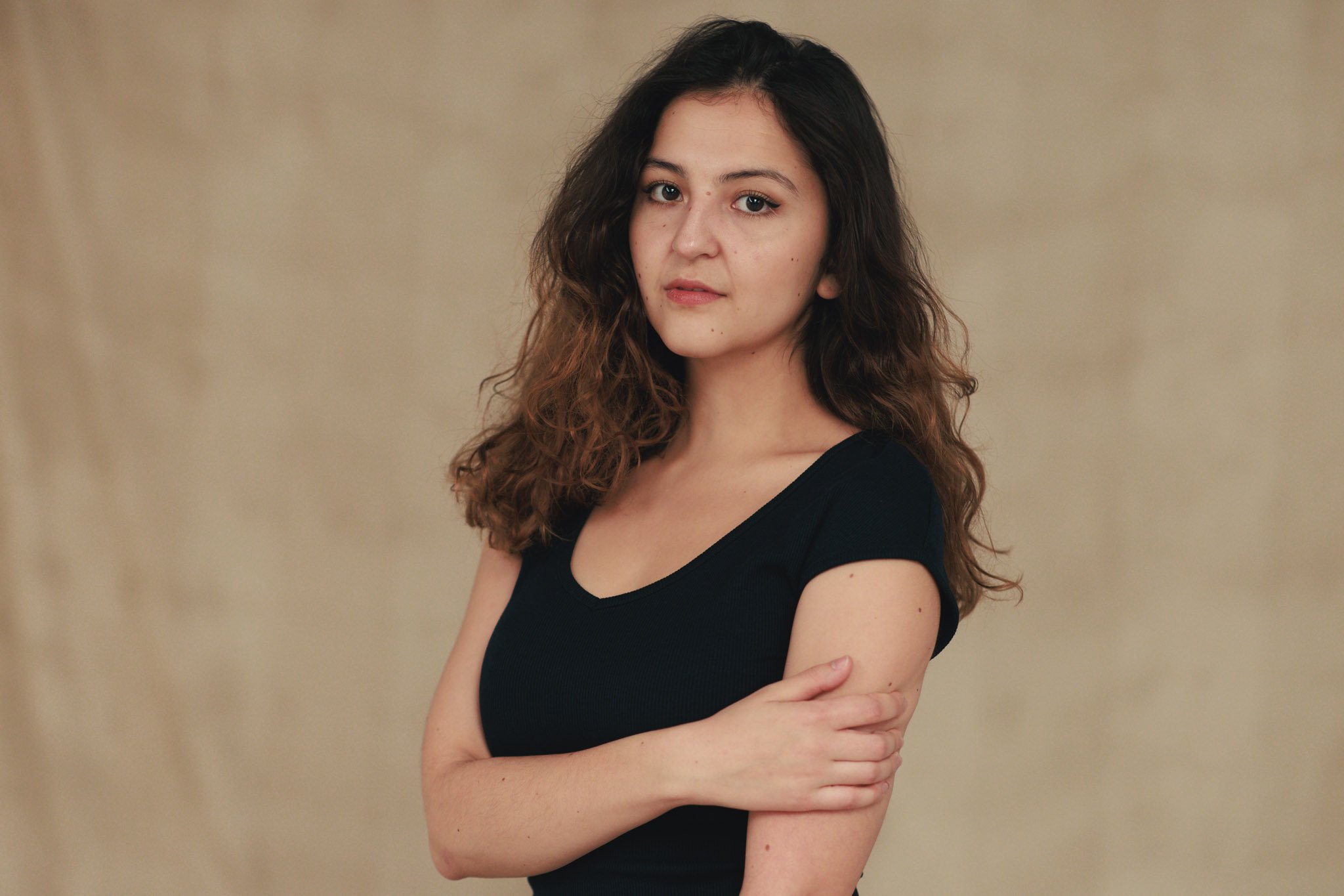 A young woman with wavy brown hair, wearing a black top, standing against a plain beige background with arms crossed.
